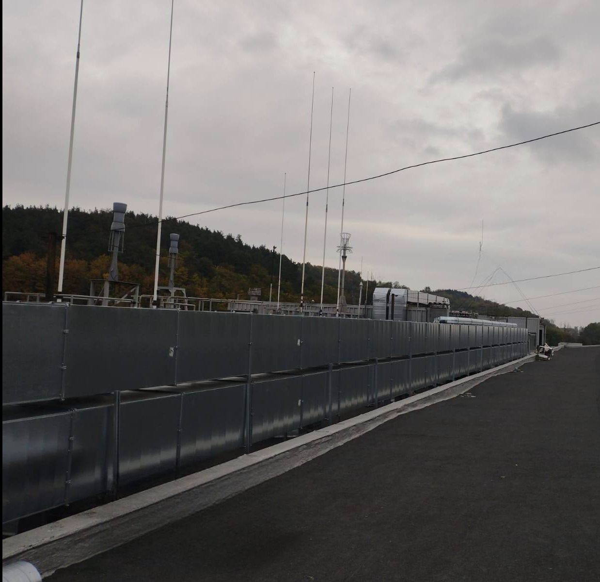 Długa instalacja wentylacyjna na dachu budynku komercyjnego, widoczne liczne wywietrzniki i anteny, pochmurne niebo w tle.