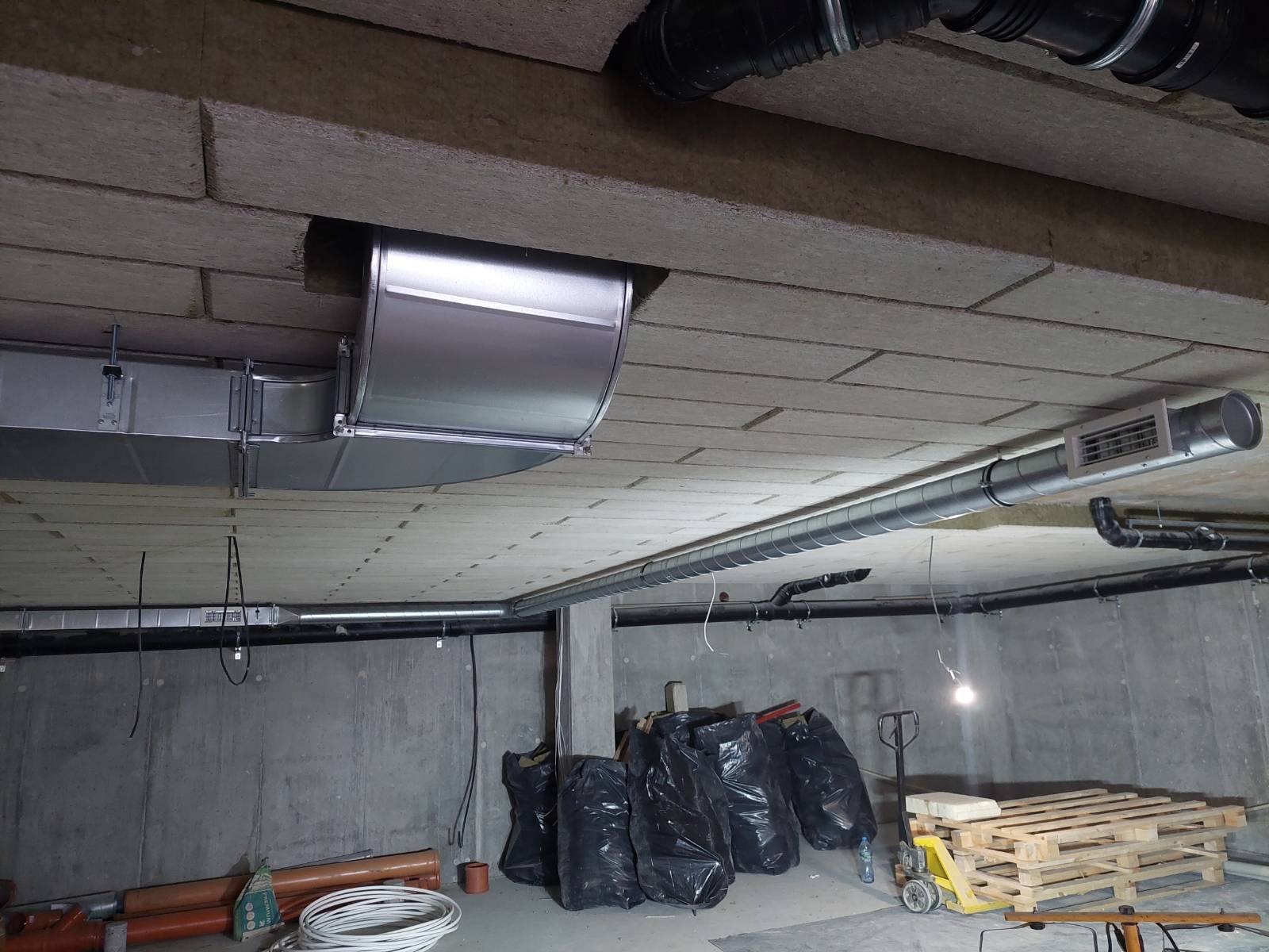 Widok na sufit w surowym stanie z zamontowanymi kanałami wentylacyjnymi, w tle czarne worki na śmieci i paleta. Widoczne elementy instalacji wentylacyjnej, rury i kształtki.