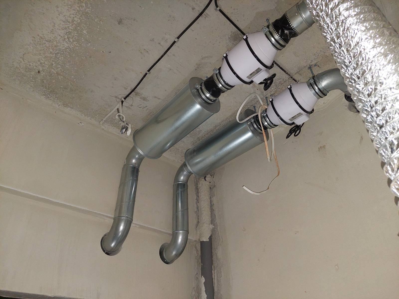 Dwa kanały wentylacyjne, jeden aluminiowy karbowany, drugi z metalu ocynkowanego, zamontowane pod betonowym sufitem, z widocznymi przewodami elektrycznymi i elementami mocującymi.