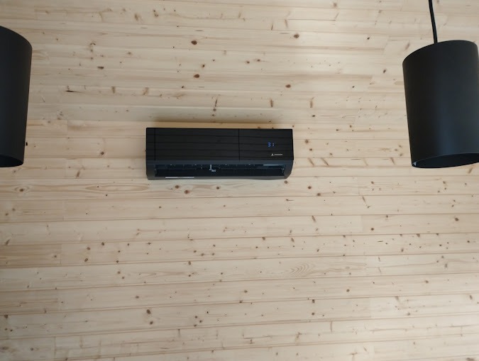 Czarny klimatyzator Mitsubishi Electric zamontowany na ścianie z jasnych, drewnianych paneli, widoczne dwie czarne lampy wiszące po bokach.