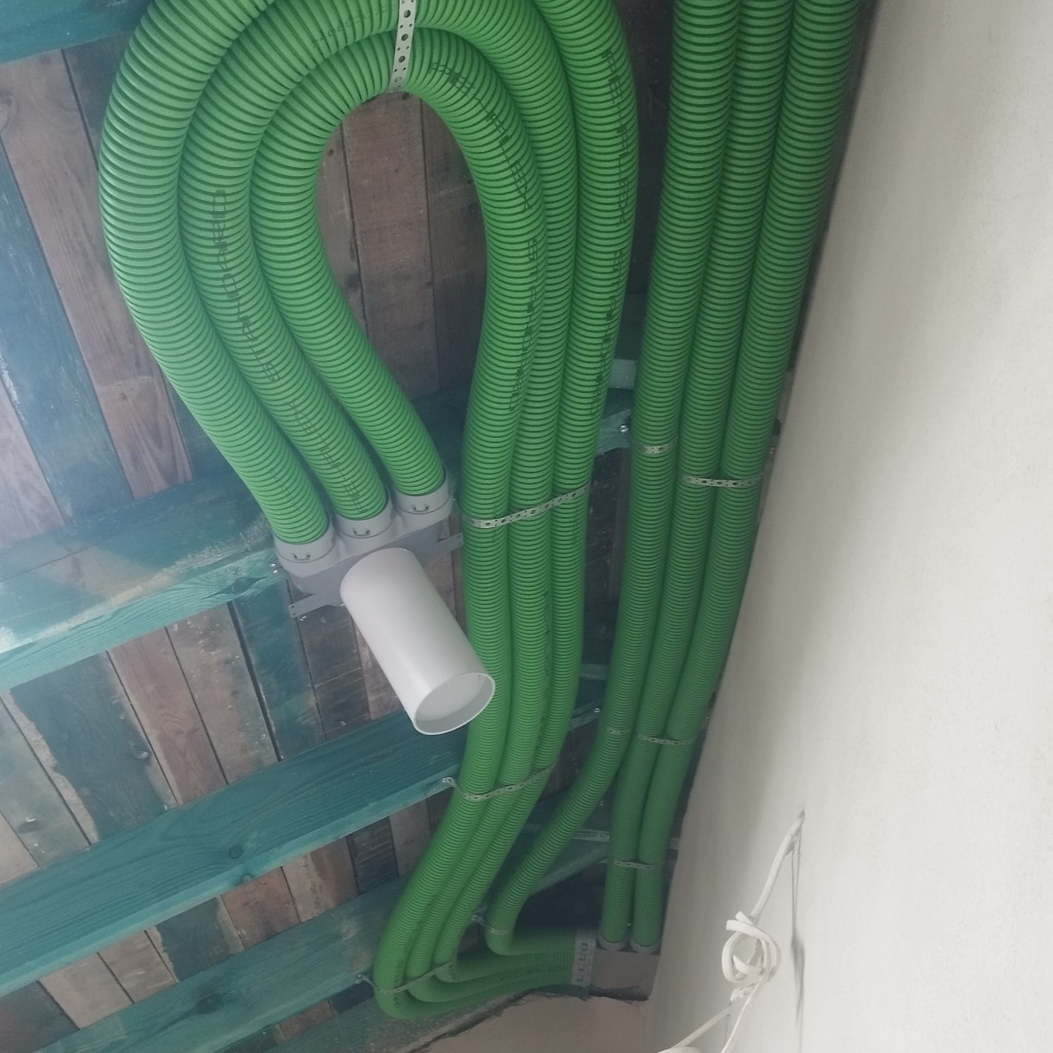 Zielone, karbowane rury wentylacyjne zamontowane pod drewnianym stropem, widoczne mocowania i biały element systemu.