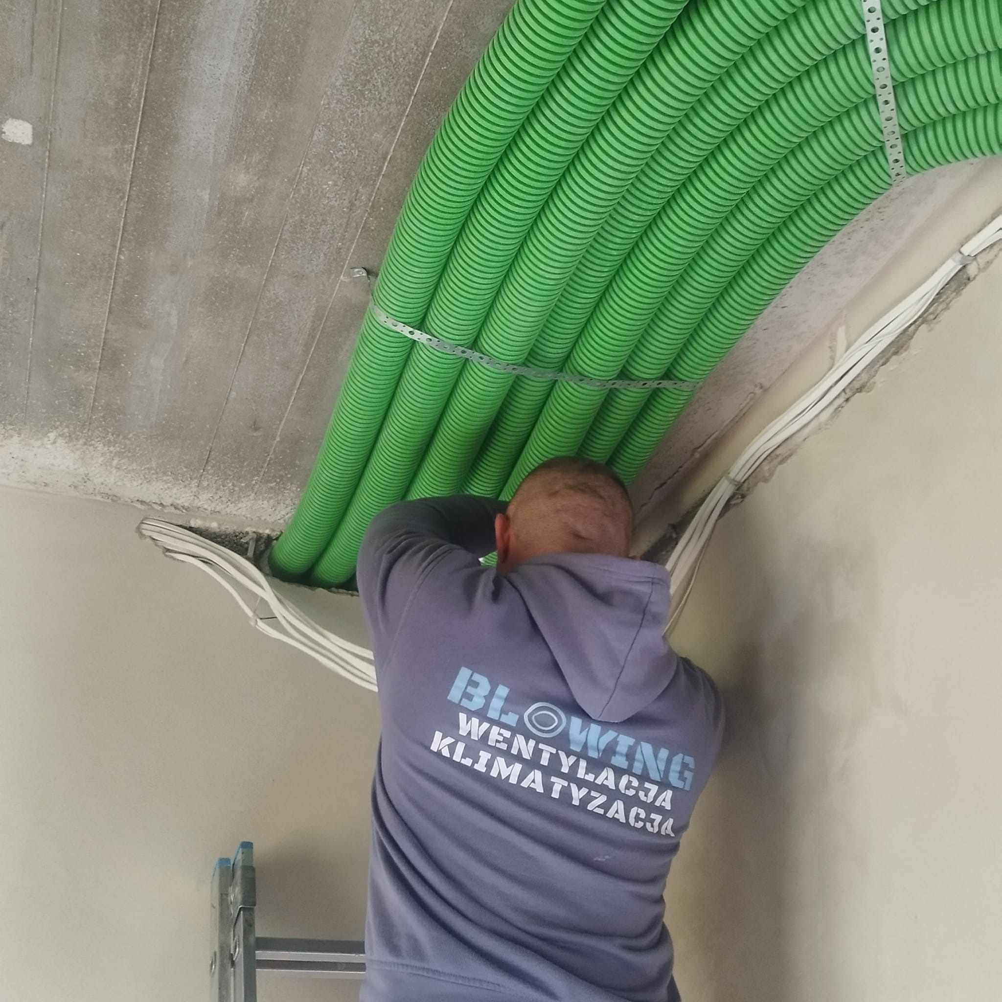 Instalator w bluzie firmowej montuje zielone rury wentylacyjne pod betonowym sufitem, widoczne metalowe uchwyty mocujące i kable elektryczne.