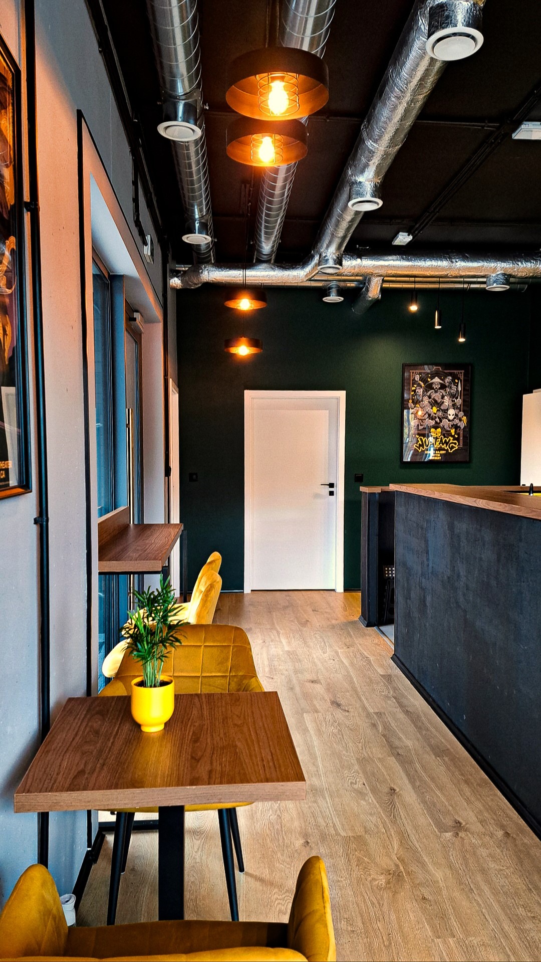 Nowoczesne wnętrze biurowe z widoczną instalacją wentylacyjną pod sufitem, oświetleniem punktowym i żółtymi akcentami kolorystycznymi w postaci krzeseł i doniczki z rośliną.