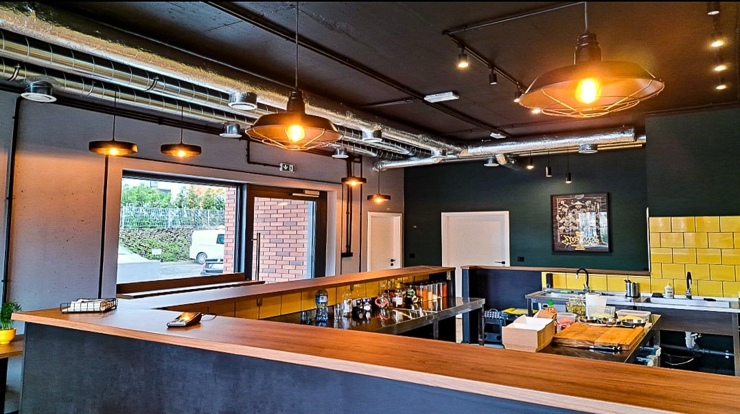 Wnętrze lokalu gastronomicznego z widocznymi rurami wentylacyjnymi pod sufitem, oświetleniem punktowym i wiszącymi lampami nad barem, oraz fragmentem instalacji wodno-kanalizacyjnej przy ścianie...