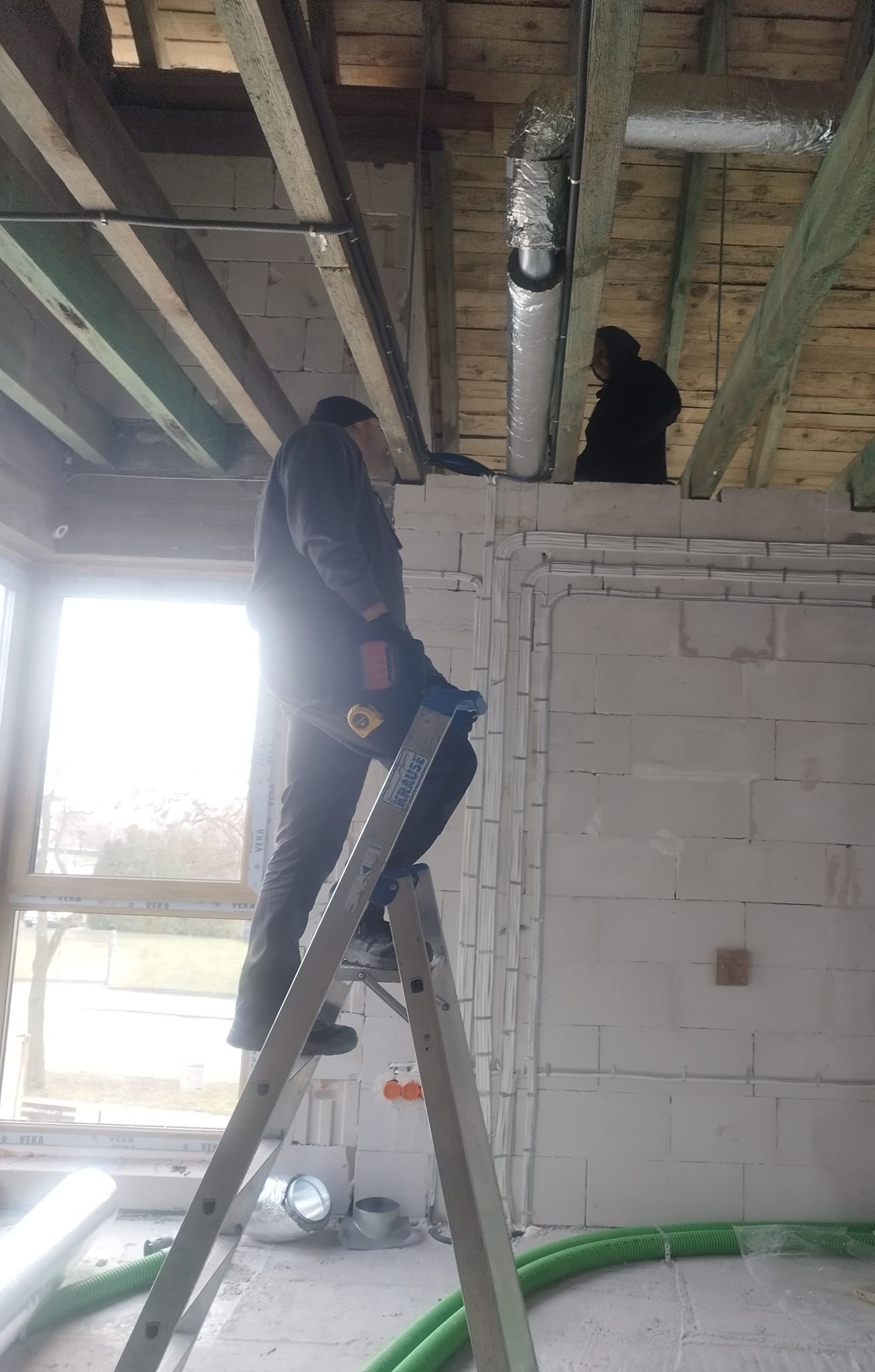 Montaż instalacji wentylacyjnej w surowym wnętrzu: monter na drabinie instaluje rurę wentylacyjną pod drewnianym stropem, widoczne przewody elektryczne i zielone rury wentylacyjne na podłodze.