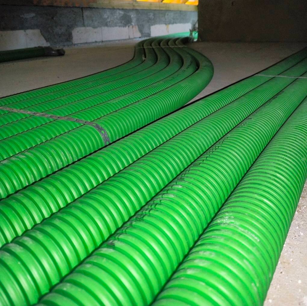 Zielone, karbowane rury wentylacyjne ułożone równolegle na podłodze w surowym pomieszczeniu z betonową podłogą i ścianami z pustaków.