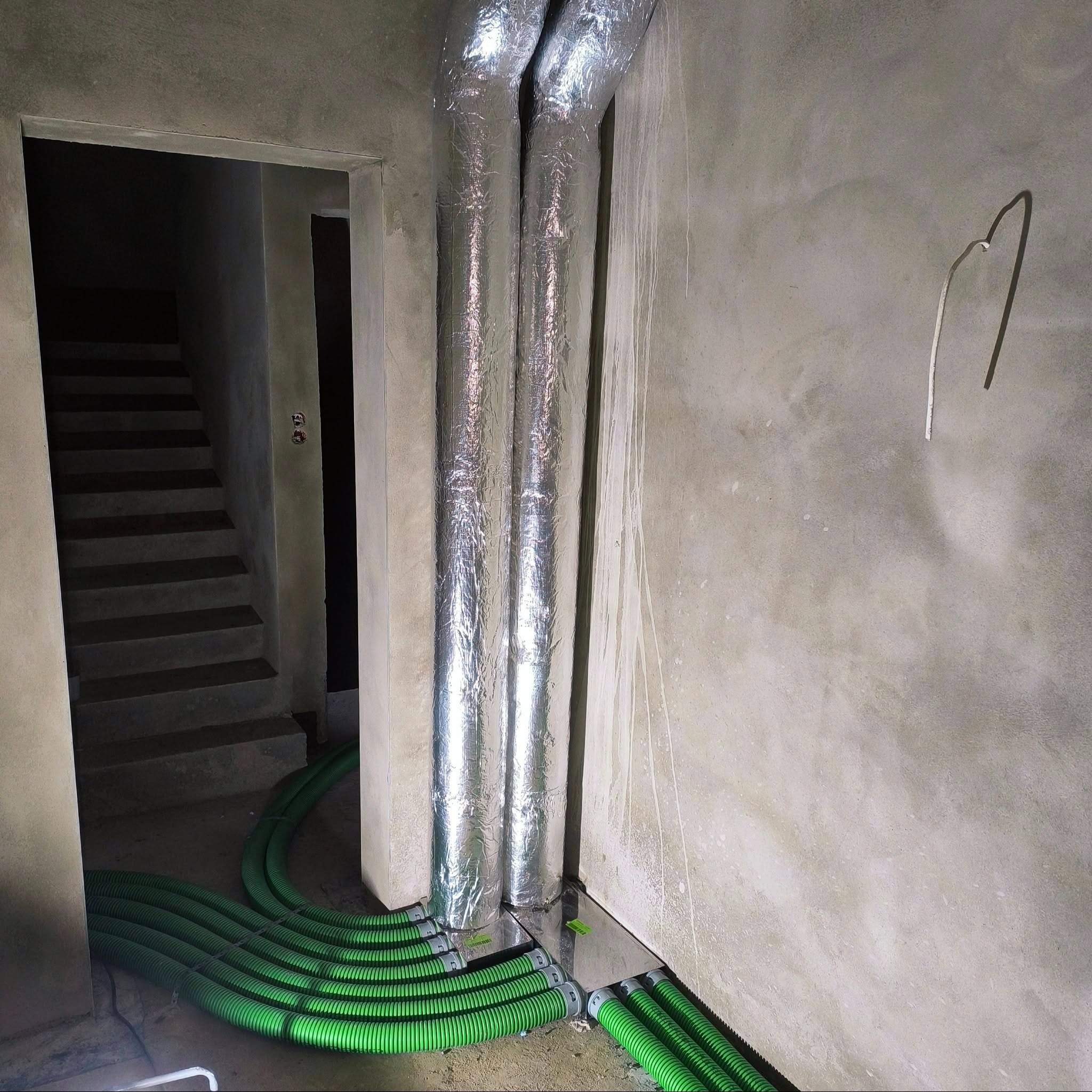 Dwa pionowe, izolowane przewody wentylacyjne przechodzące przez podłogę, z podłączonymi zielonymi, elastycznymi rurami wentylacyjnymi, w surowym pomieszczeniu ze schodami w tle.