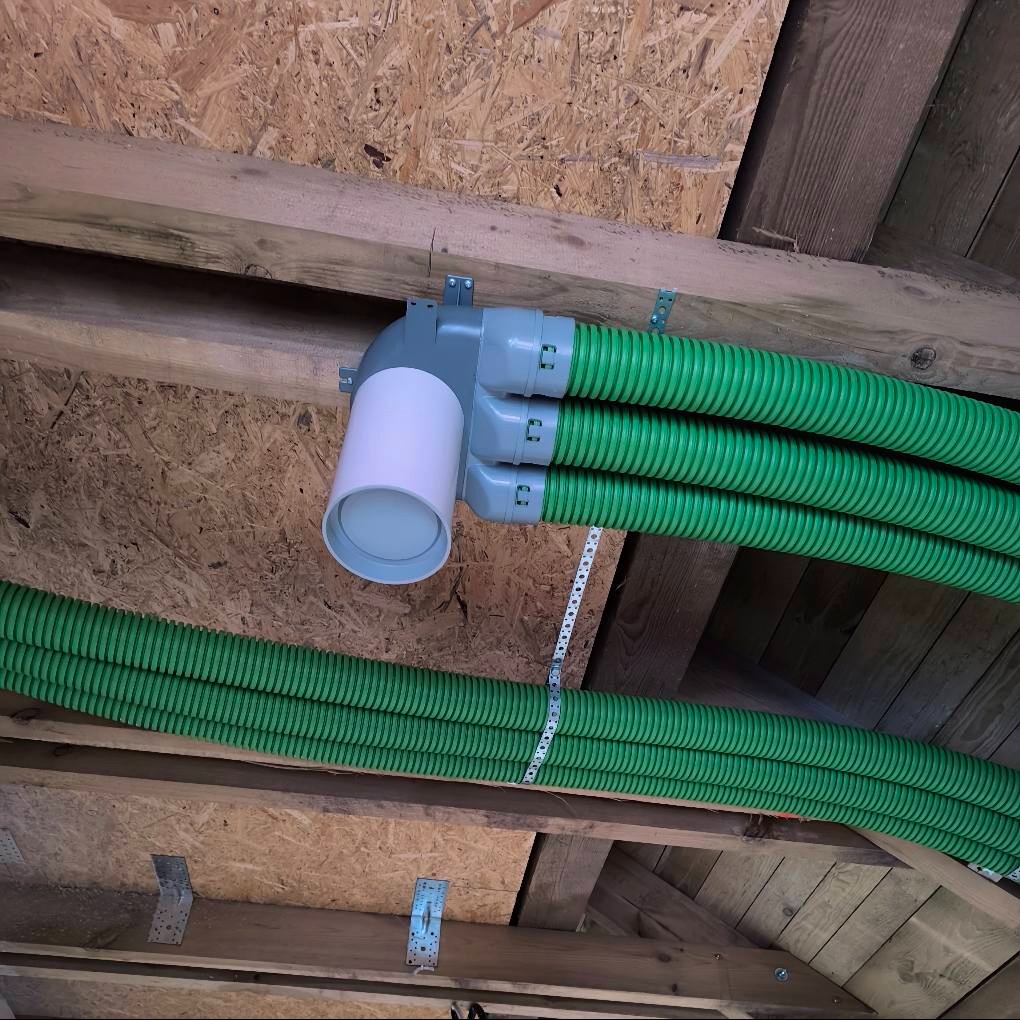 Zielone, karbowane rury wentylacyjne przymocowane do drewnianej konstrukcji stropu za pomocą metalowych uchwytów i taśmy perforowanej. Szara kształtka rozdzielająca rury z białym, cylindrycznym...