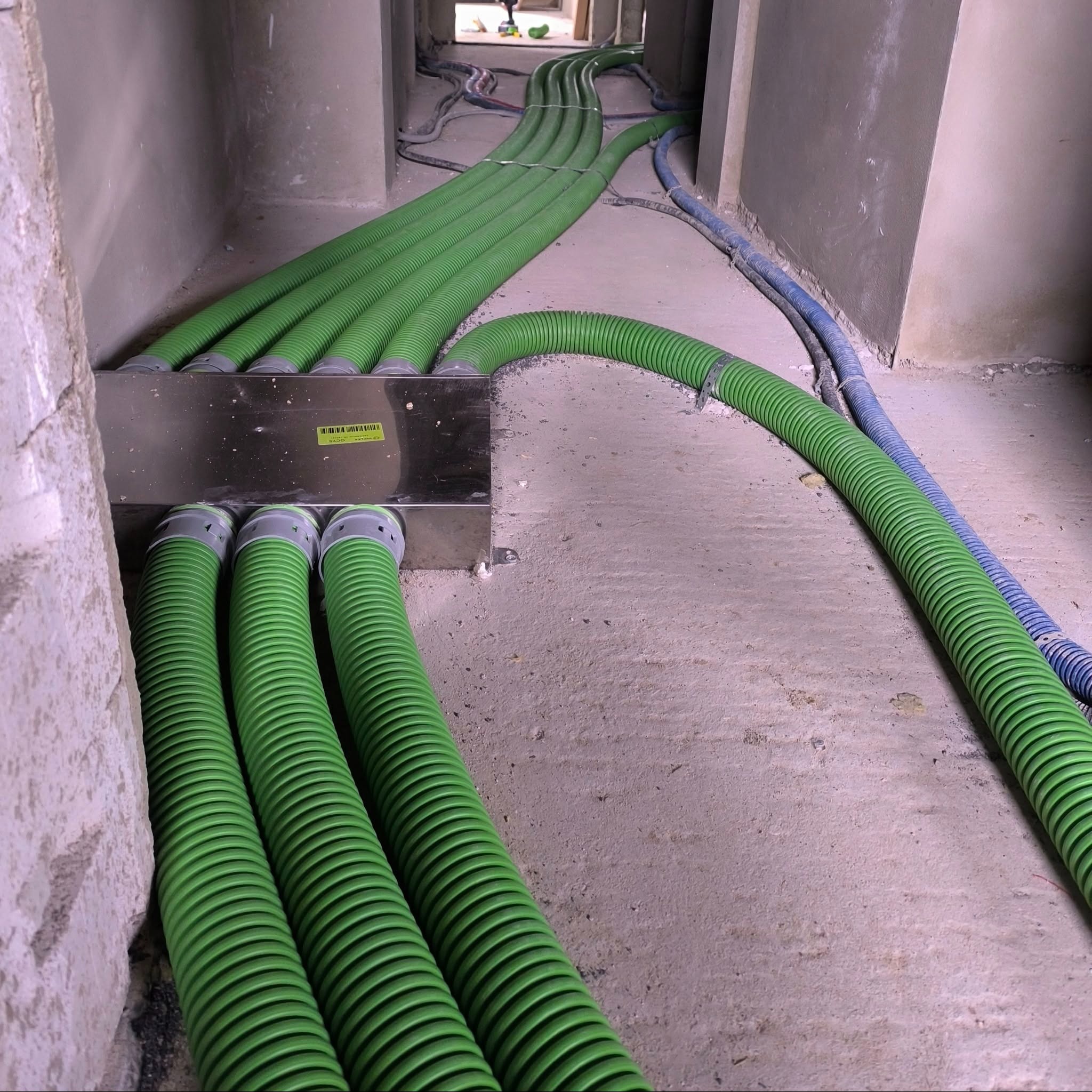 Wnętrze w trakcie prac instalacyjnych: zielone, karbowane rury wentylacyjne wychodzące z metalowej skrzynki, ułożone wzdłuż surowej ściany i podłogi.