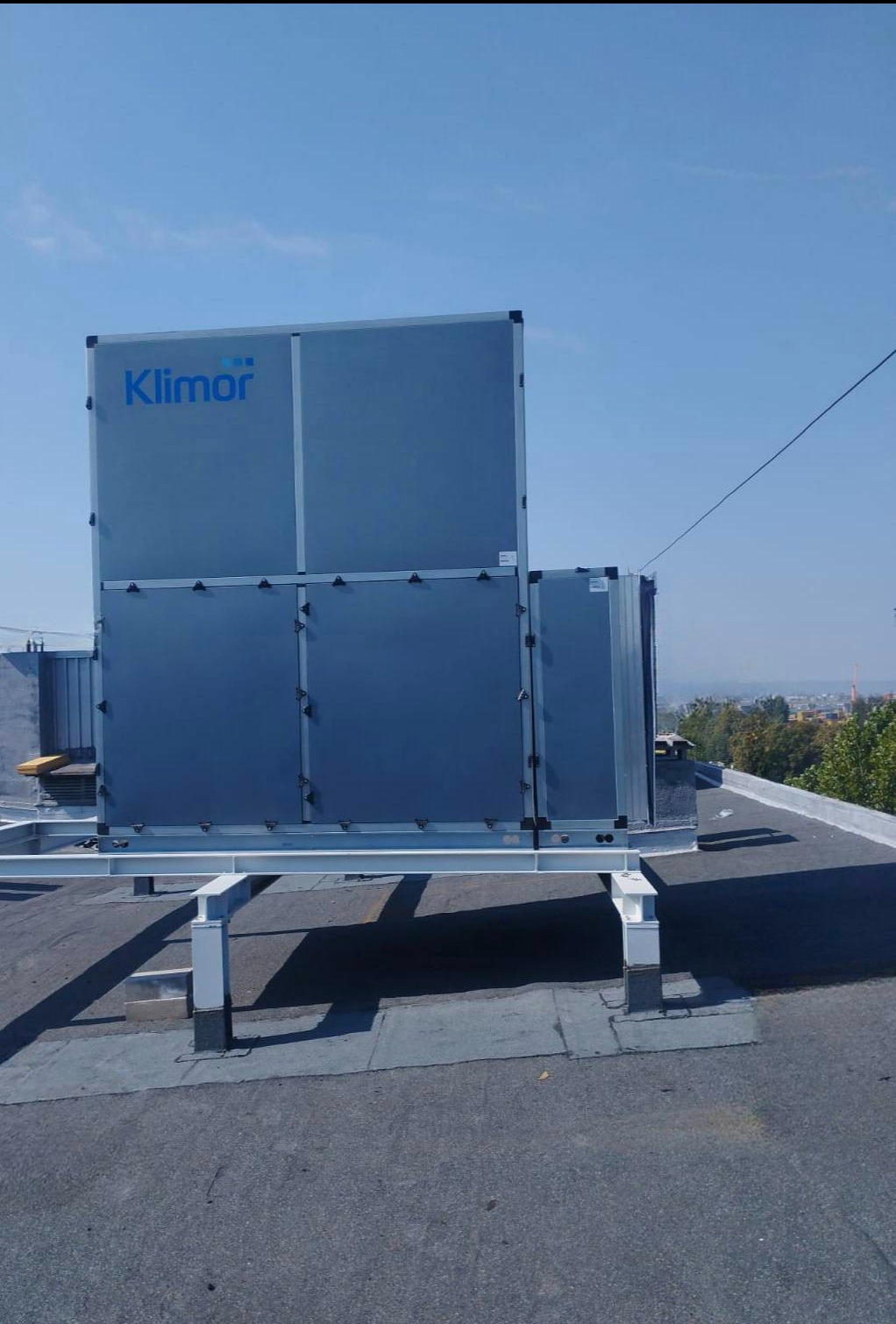 Centrala wentylacyjna Klimor zamontowana na dachu budynku, widok z dołu. Metalowa konstrukcja wsporcza. W tle błękitne niebo i panorama miasta.