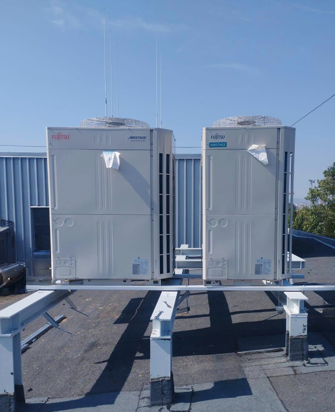 Dwa urządzenia klimatyzacyjne Fujitsu Airstage na dachu, zamontowane na białej konstrukcji wsporczej. Widoczne anteny i elementy instalacji na tle błękitnego nieba.