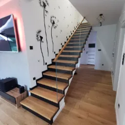 Nowoczesne schody drewniane z balustradą szklaną, czarne stopnice, drewniany parkiet, ściana z motywem dmuchawców.