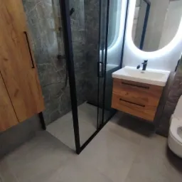 Nowoczesna łazienka z kabiną prysznicową typu walk-in z czarnymi profilami, drewnianą szafką pod umywalką z okrągłym lustrem podświetlonym LED i szarą marmurową okładziną ścian.