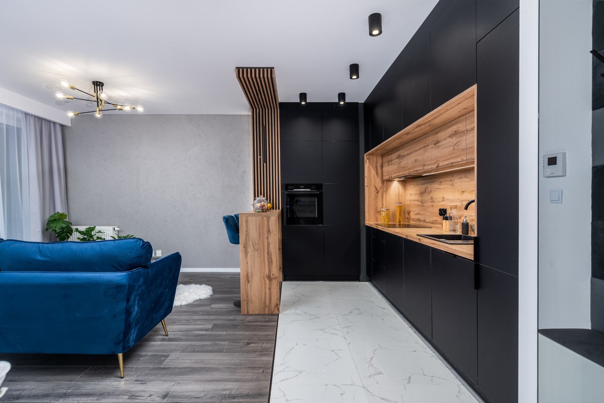 Nowoczesne wnętrze z czarną zabudową kuchenną z drewnianymi akcentami i niebieską sofą w Krakowie, widok na salon z designerską lampą.