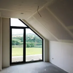 Poddasze z oknem szczytowym w trakcie ocieplania, widoczne płyty karton-gips na skosach i suficie, wstępnie zaszpachlowane łączenia płyt, wystający kabel elektryczny.