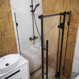 Nowoczesna łazienka z drewnopodobną mozaiką na ścianie, białą umywalką nablatową na szafce z marmurowym blatem i kabiną prysznicową z czarnymi profilami.