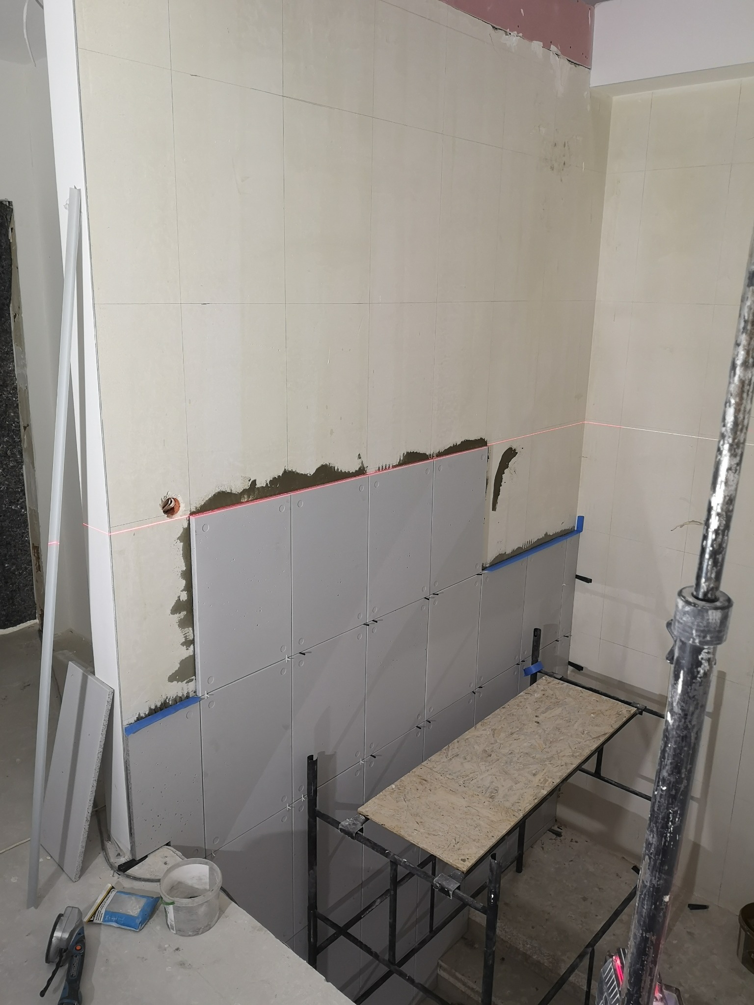Montaż szarych płyt imitujących beton na ścianie, widoczna poziomica laserowa, rusztowanie i narzędzia glazurnicze.
