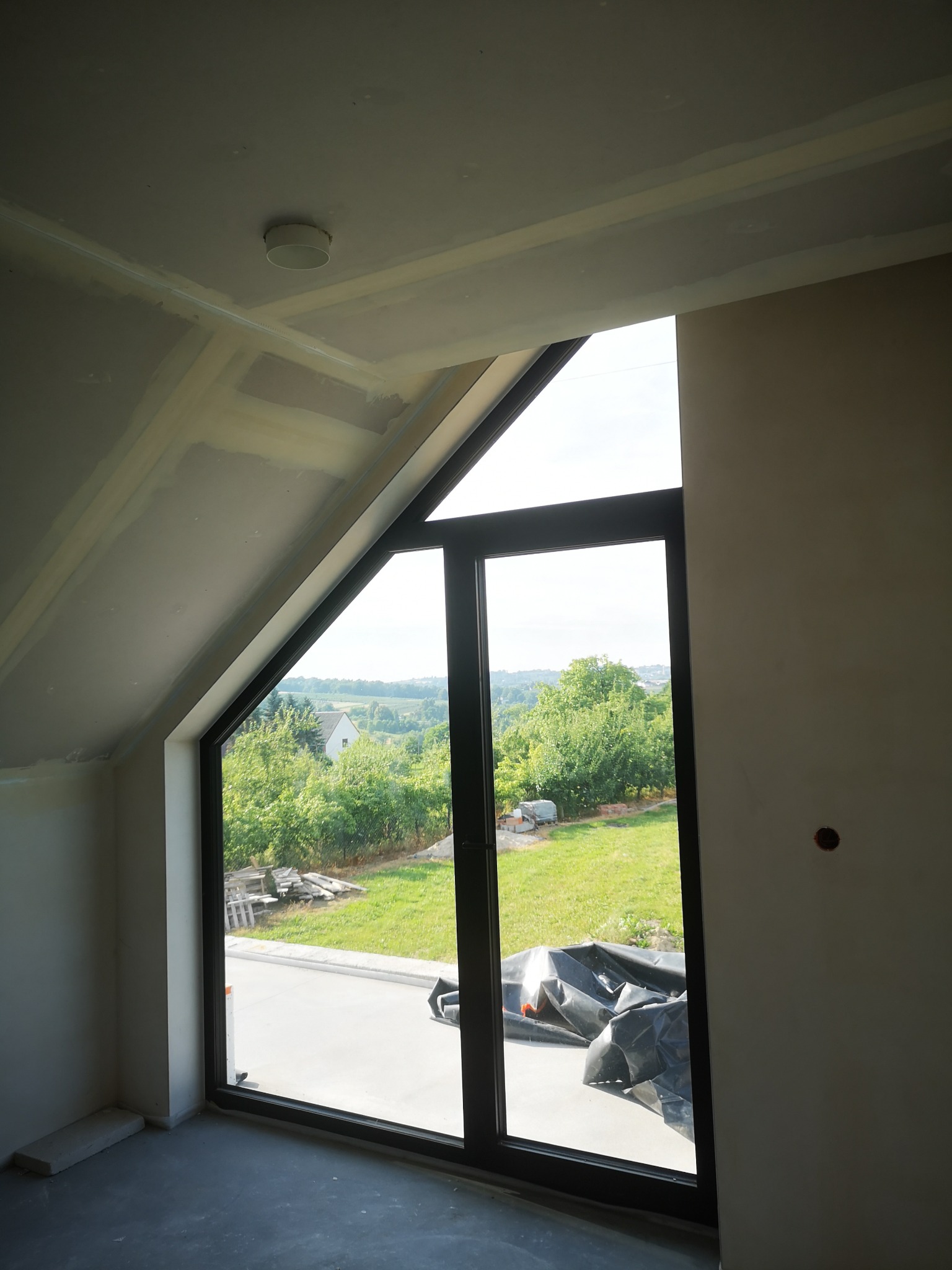 Poddasze z oknem szczytowym w trakcie prac wykończeniowych, widoczne łączenia płyt gipsowo-kartonowych, szpachlowane i przygotowane do dalszych prac. Na zewnątrz widok na zielony ogród i zabudowę.