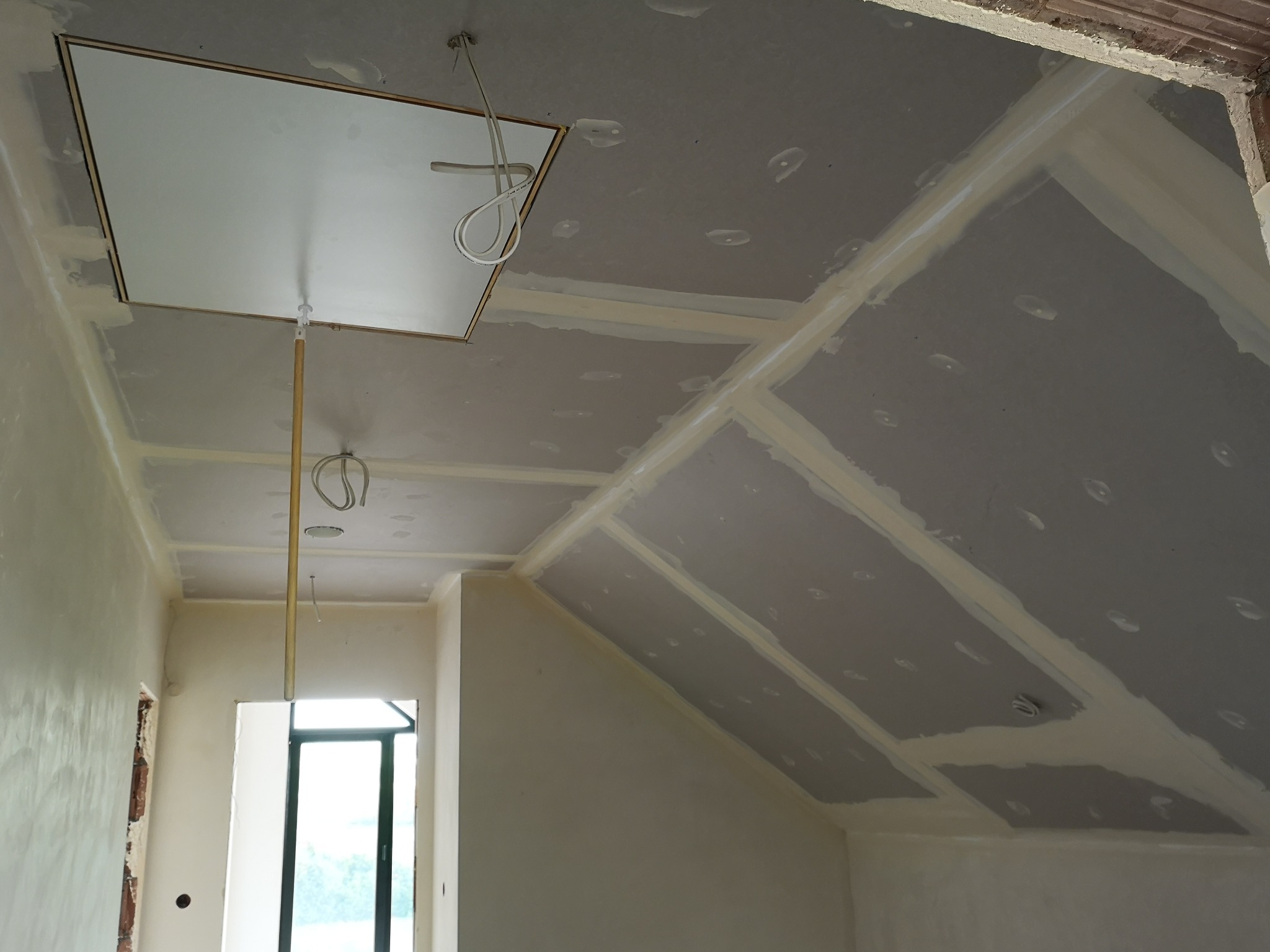 Poddasze w trakcie prac wykończeniowych: widoczne płyty kartonowo-gipsowe na skośnym suficie, łączenia wzmocnione taśmą, otwór rewizyjny z widoczną instalacją elektryczną, ściany przygotowane...