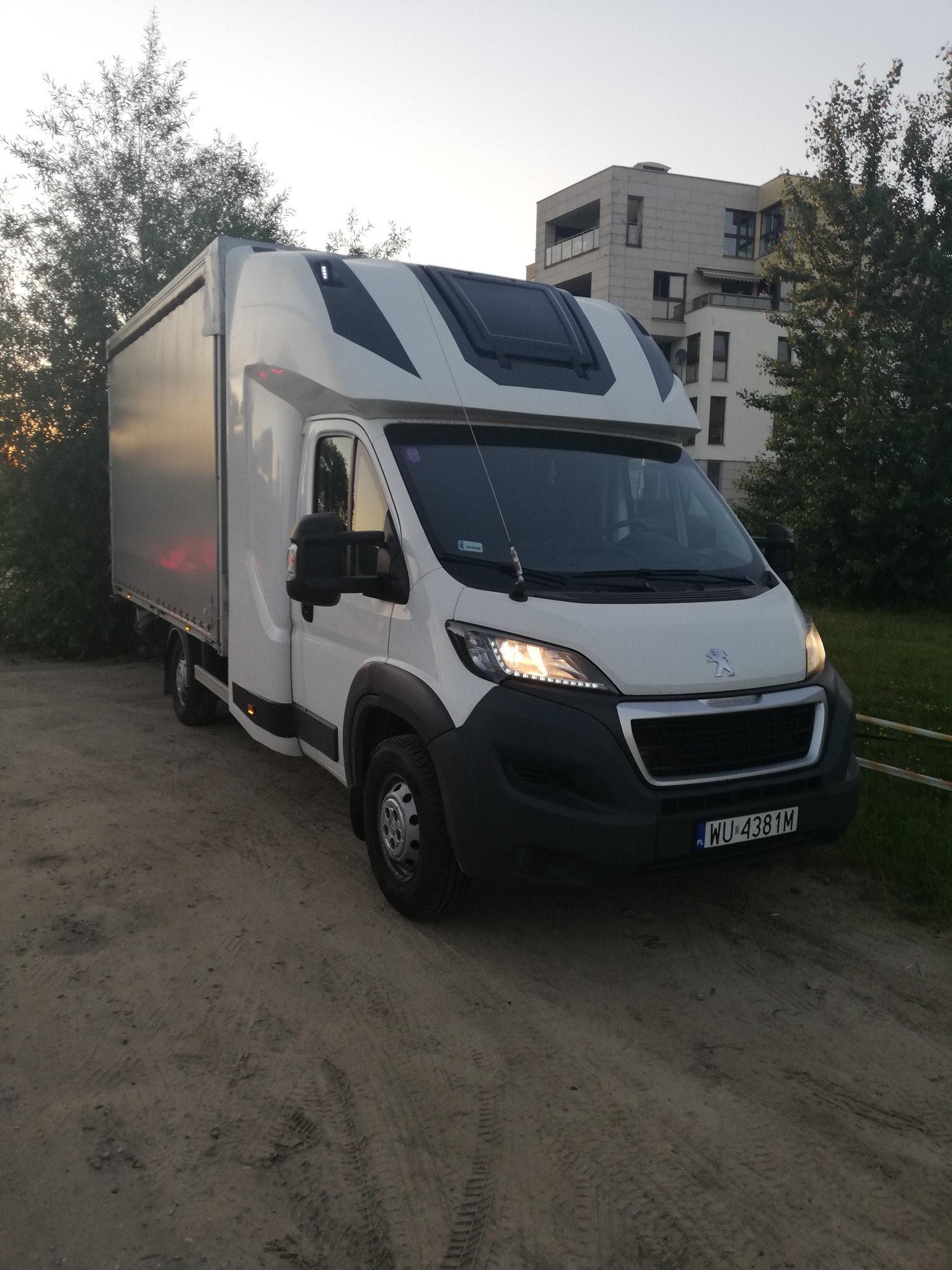 Biały bus dostawczy Peugeot z zabudową kontenerową, zaparkowany na ziemnej drodze, z widoczną tablicą rejestracyjną i fragmentem nowoczesnego budynku w tle, sfotografowany o zmierzchu.