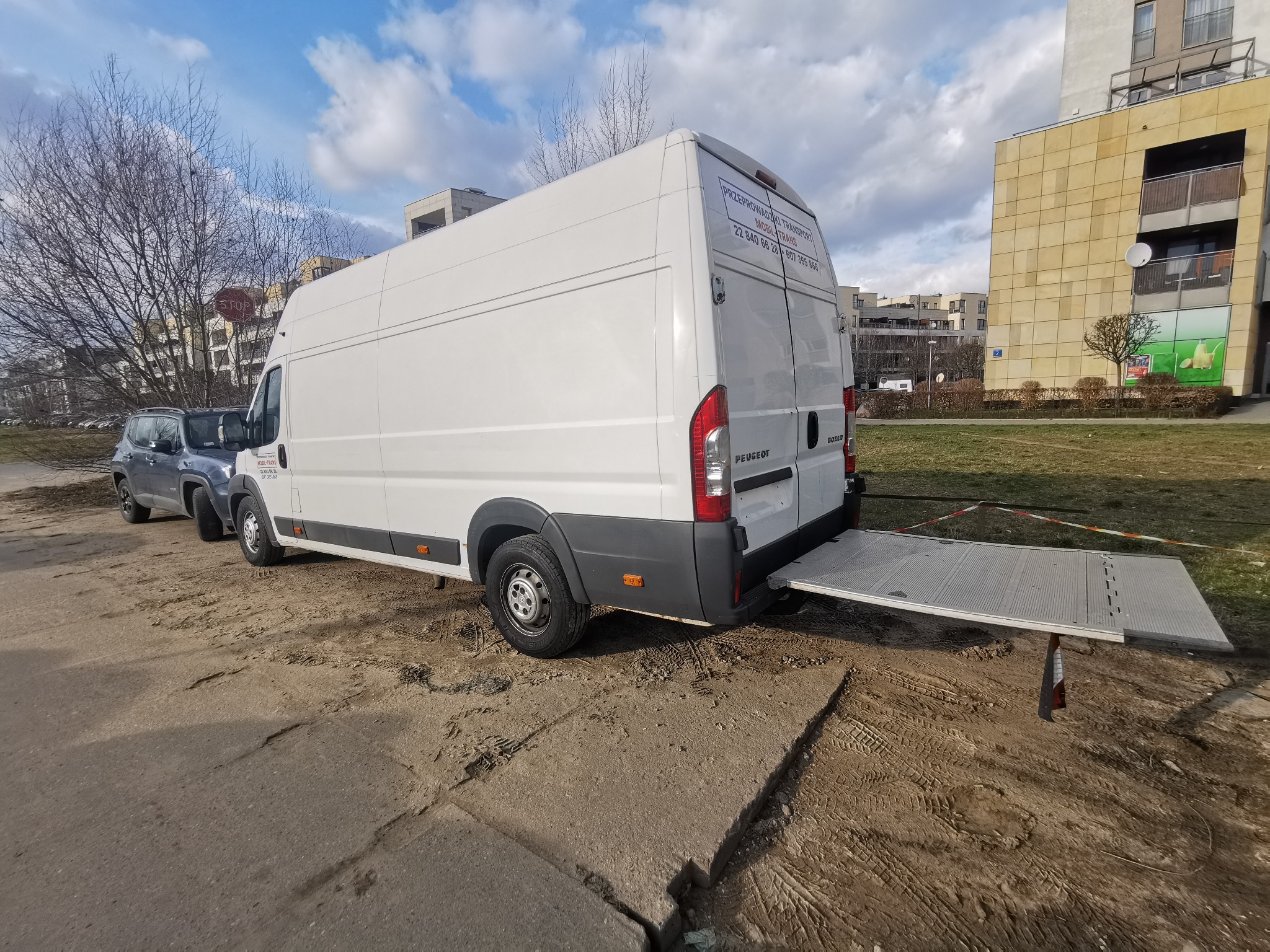 Biały bus marki Peugeot z rozłożoną rampą załadunkową na betonowym podłożu z widocznymi śladami opon, zaparkowany w miejskiej scenerii z budynkami w tle.