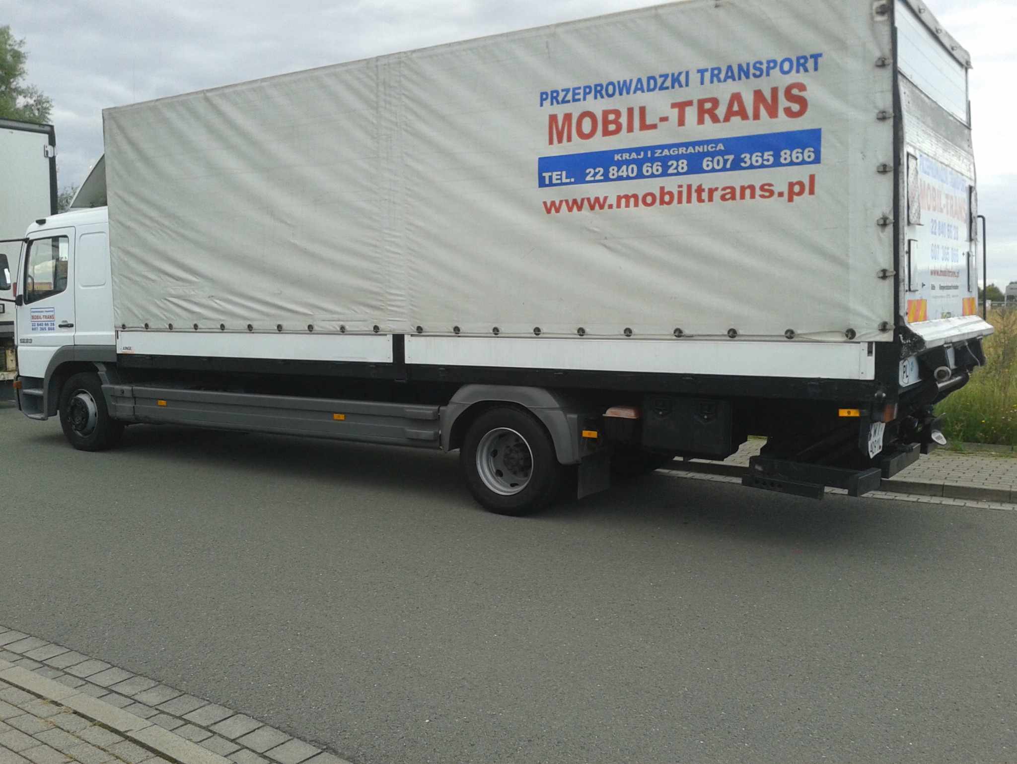 Biała ciężarówka z plandeką firmy transportowej Mobil-Trans, z widocznym numerem telefonu i adresem strony internetowej, zaparkowana na szarej nawierzchni asfaltowej.