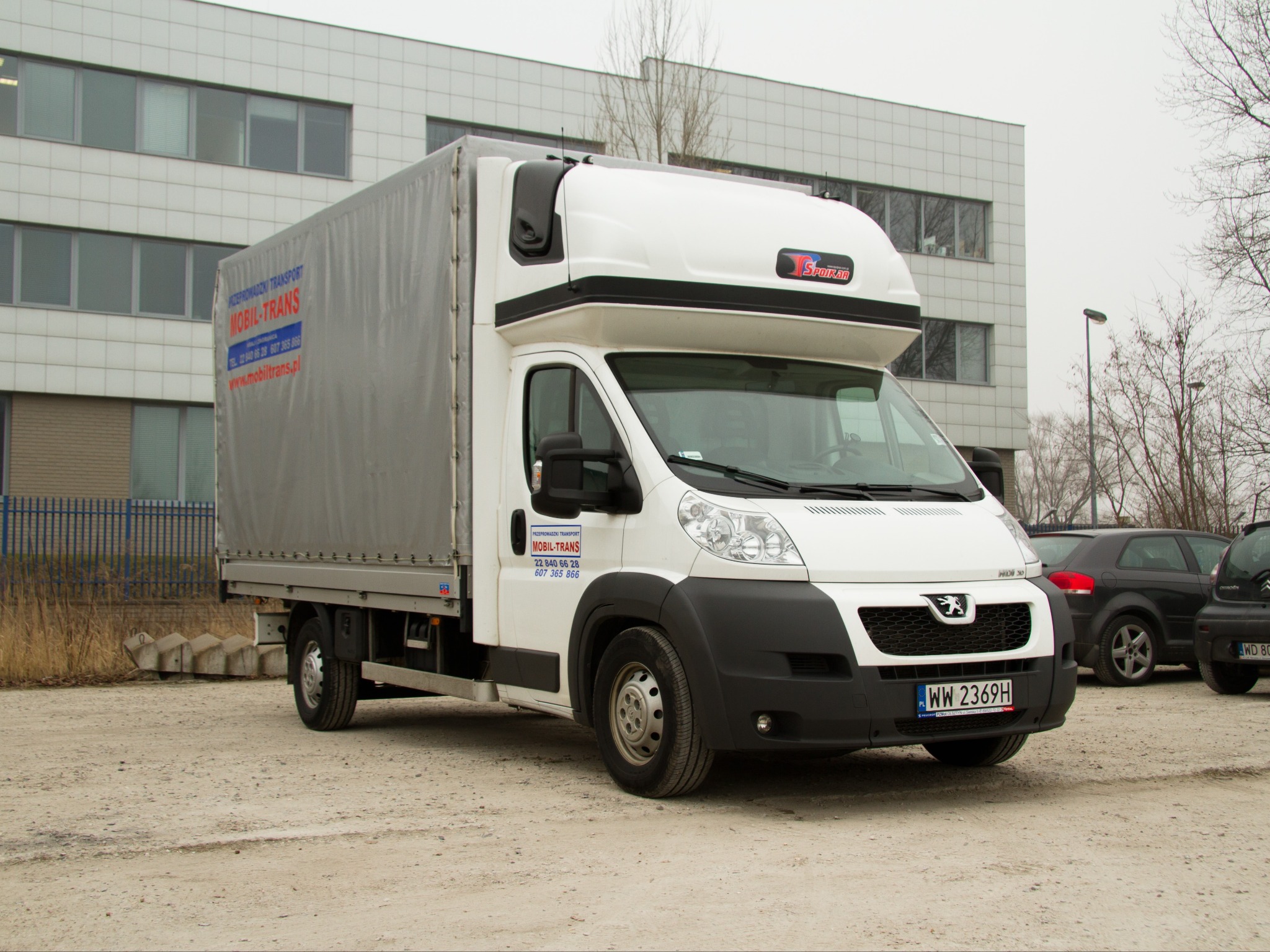 Biały bus dostawczy z szarą plandeką i logo firmy przewozowej Mobil-Trans na tle biurowca i zaparkowanych samochodów.