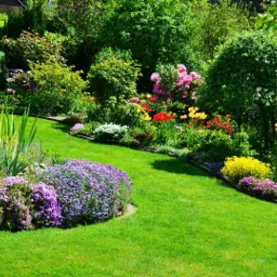 Starannie utrzymany ogr&oacute;d z intensywnie zielonym trawnikiem, kolorowymi rabatami kwiatowymi i formowanymi krzewami, otoczony bujną zielenią drzew.