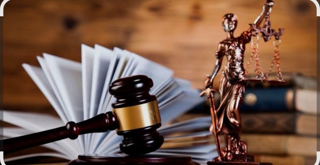 Temida, młotek sędziowski i otwarte akta na drewnianym tle. Symbol sprawiedliwości i prawa w ciepłej tonacji.