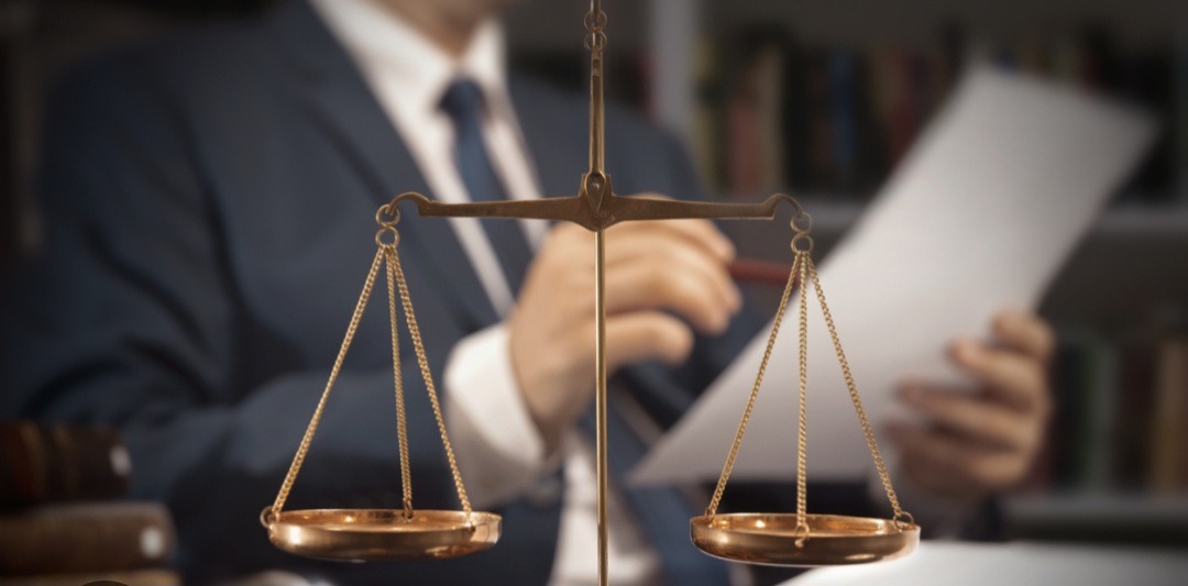 Złota waga sprawiedliwości na pierwszym planie, w tle prawnik w garniturze analizujący dokumenty w gabinecie z biblioteczką. Symbol prawa i sprawiedliwości.