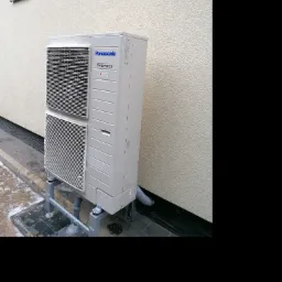 Zewnętrzna jednostka klimatyzacji Panasonic Inverter, zamontowana na metalowej konstrukcji przy ścianie budynku z widocznymi małymi oknami, na częściowo oblodzonej kostce brukowej.