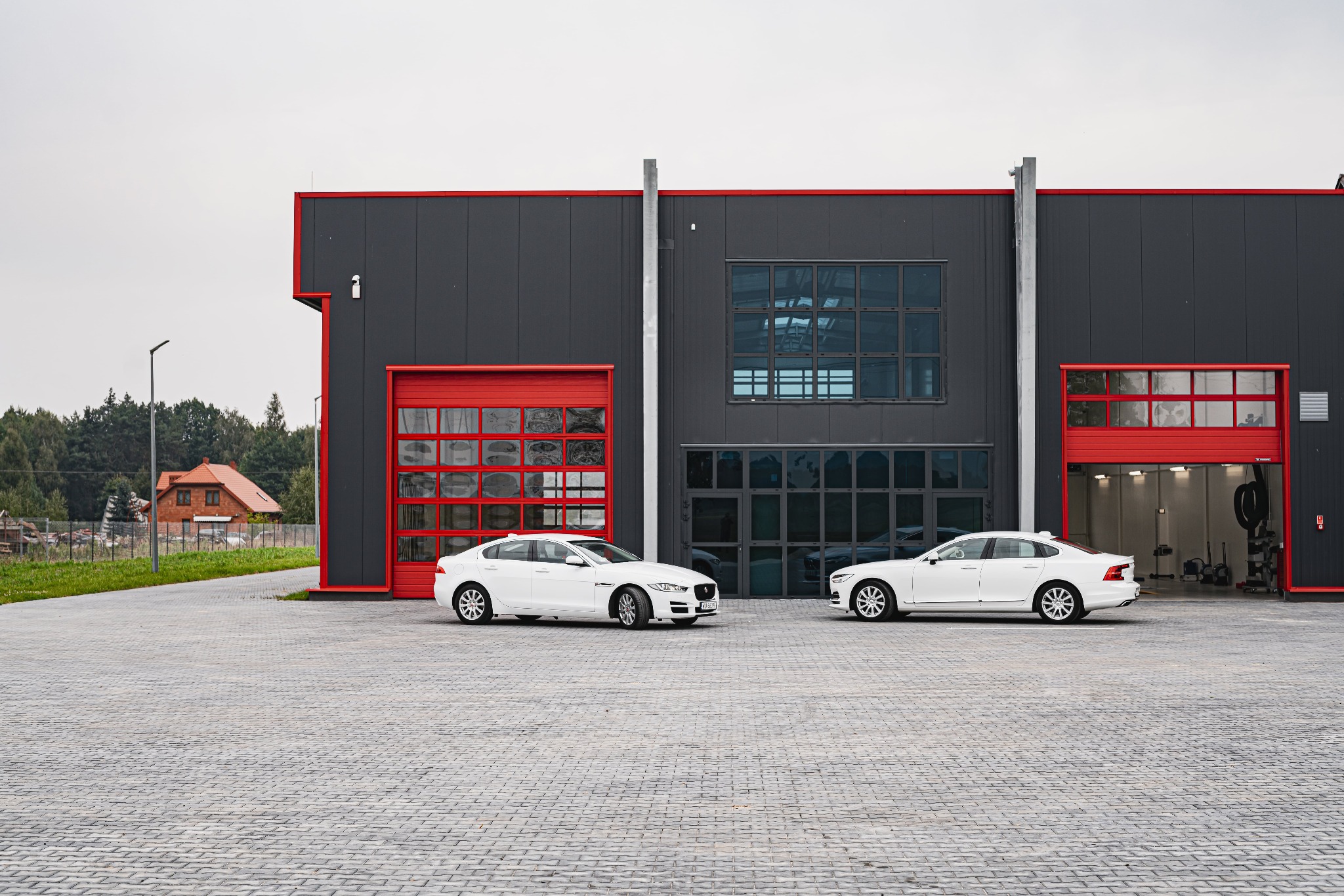 Dwa białe samochody zaparkowane przed nowoczesnym budynkiem z szarymi ścianami i czerwonymi bramami garażowymi.