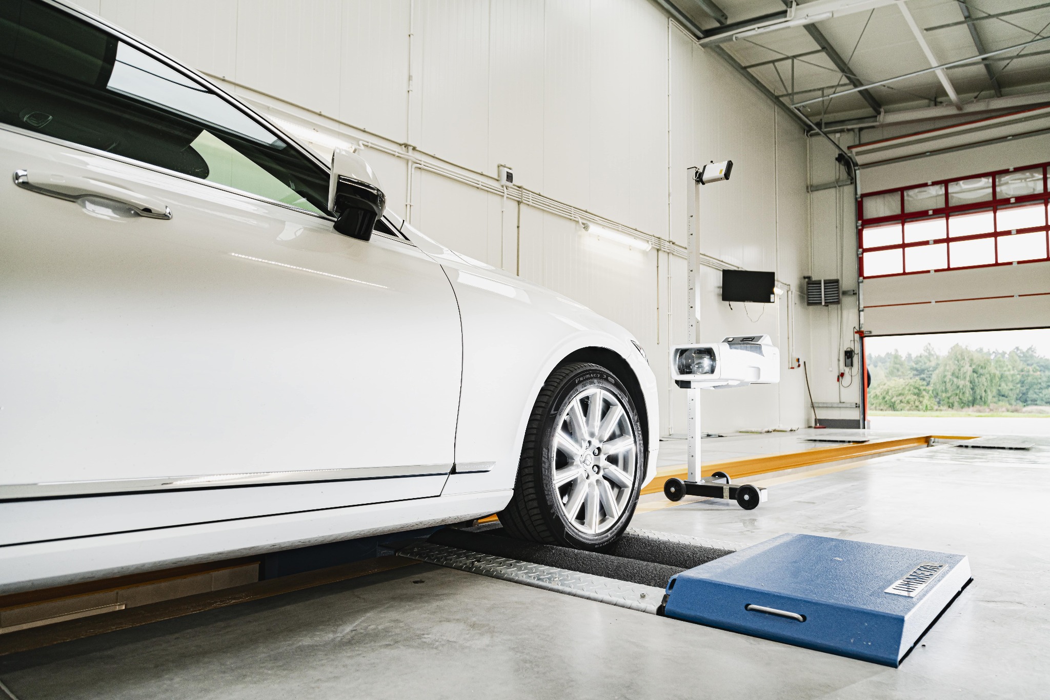 Biały samochód osobowy na stanowisku do geometrii kół w jasnym warsztacie, widoczny sprzęt diagnostyczny i otwarta brama garażowa.