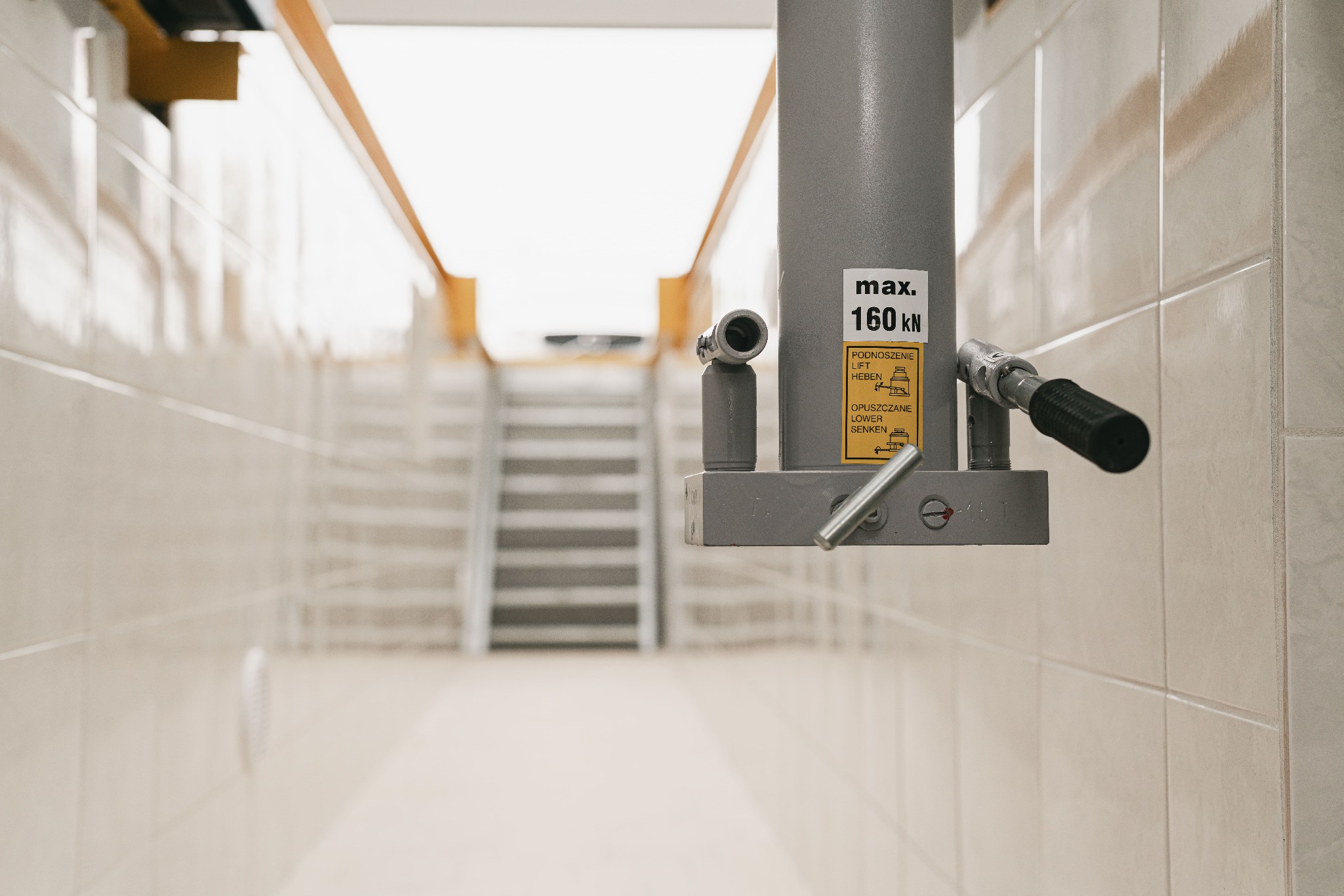 Hydrauliczny podnośnik kolumnowy z napisem 'max. 160 kN' w jasnym pomieszczeniu wyłożonym kafelkami, widoczne schody w tle.