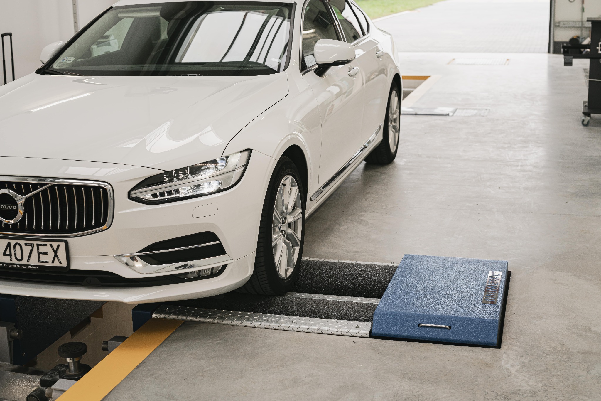 Biały samochód Volvo na podnośniku diagnostycznym w warsztacie, widoczny fragment tablicy rejestracyjnej i logo Volvo na grillu.