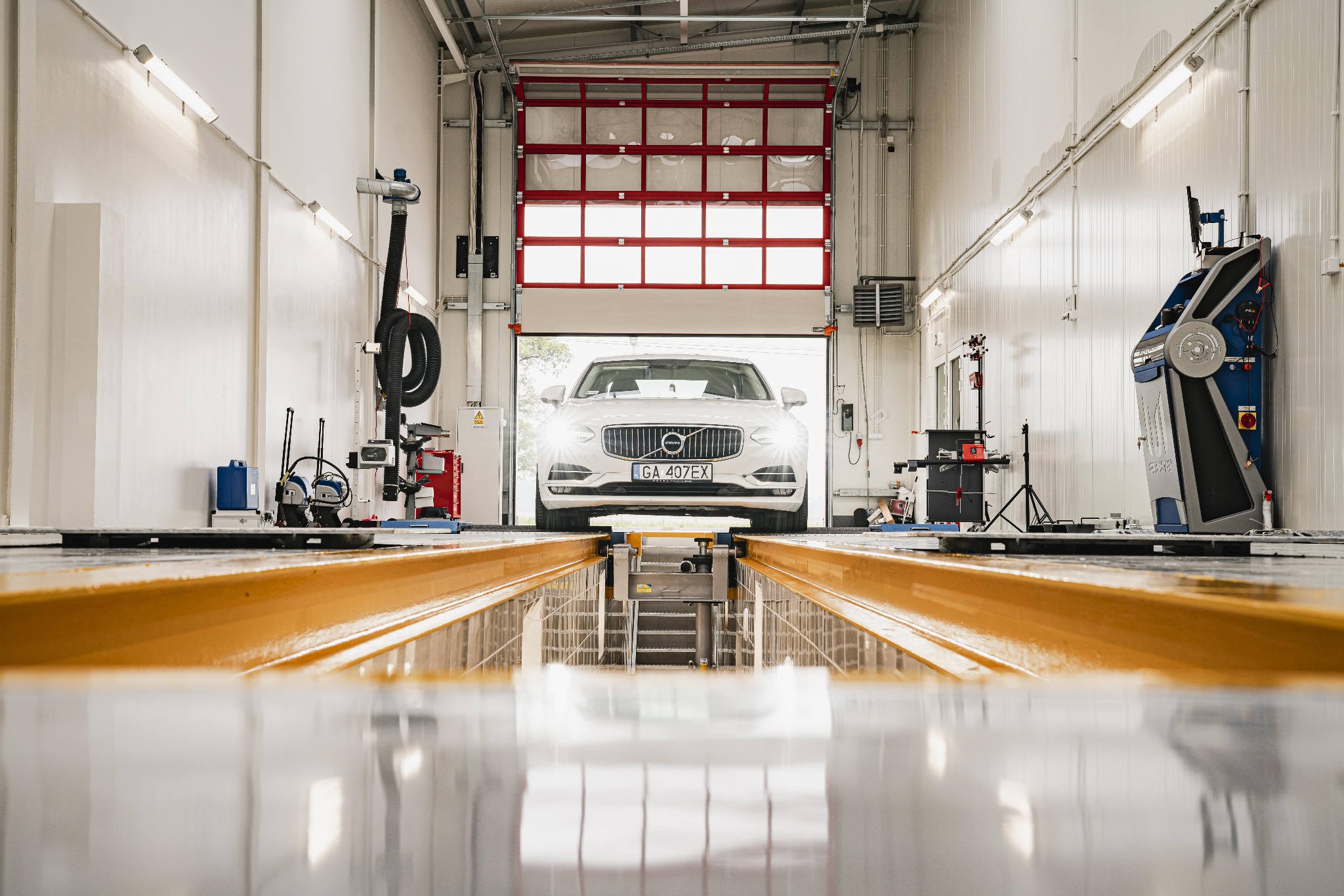 Biały samochód marki Volvo na podnośniku w nowoczesnym, jasnym warsztacie z czerwono-białą bramą garażową w tle.