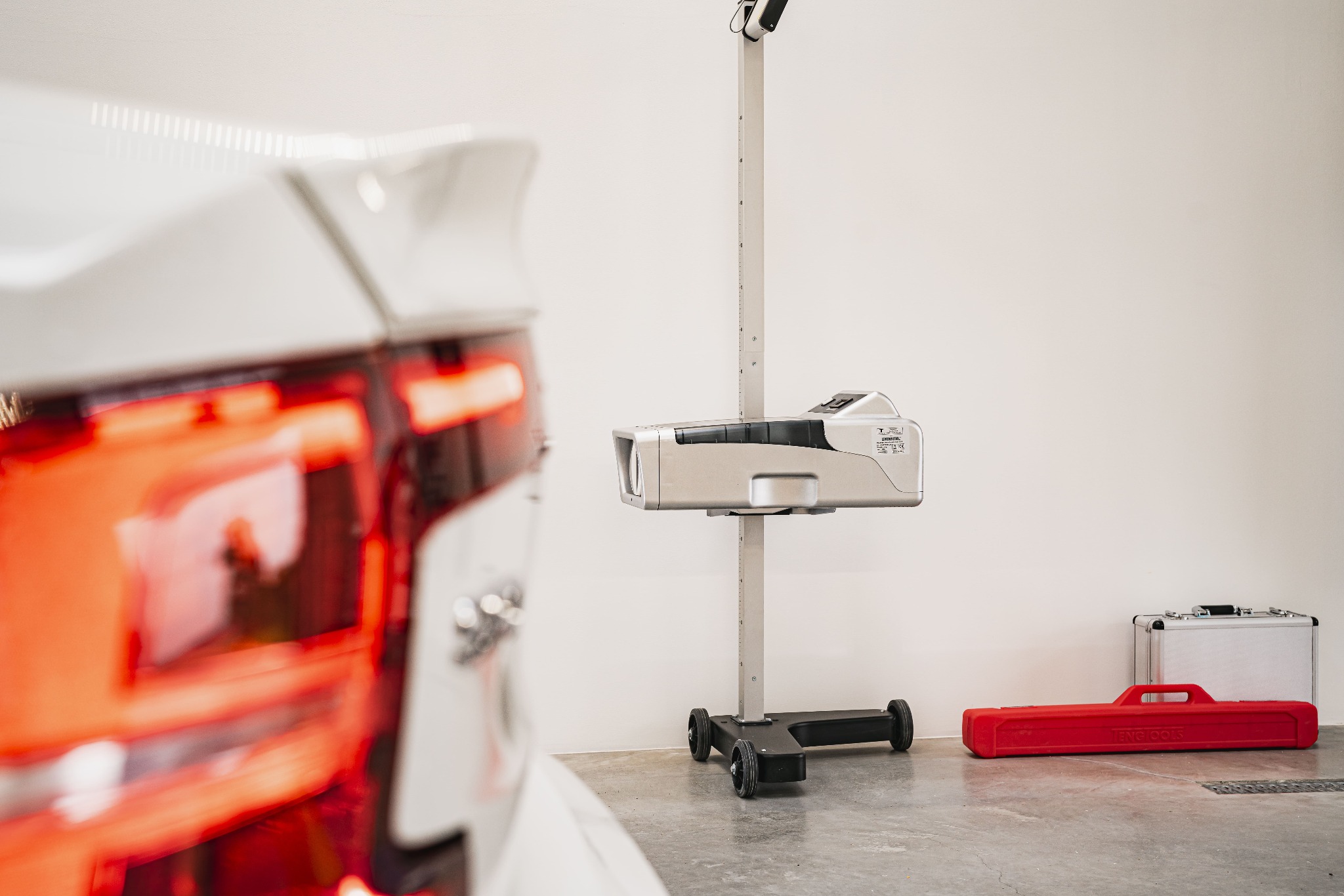 Urządzenie do regulacji świateł samochodowych w warsztacie, widoczny fragment tylnej lampy białego pojazdu, czerwona skrzynka narzędziowa i aluminiowa walizka.