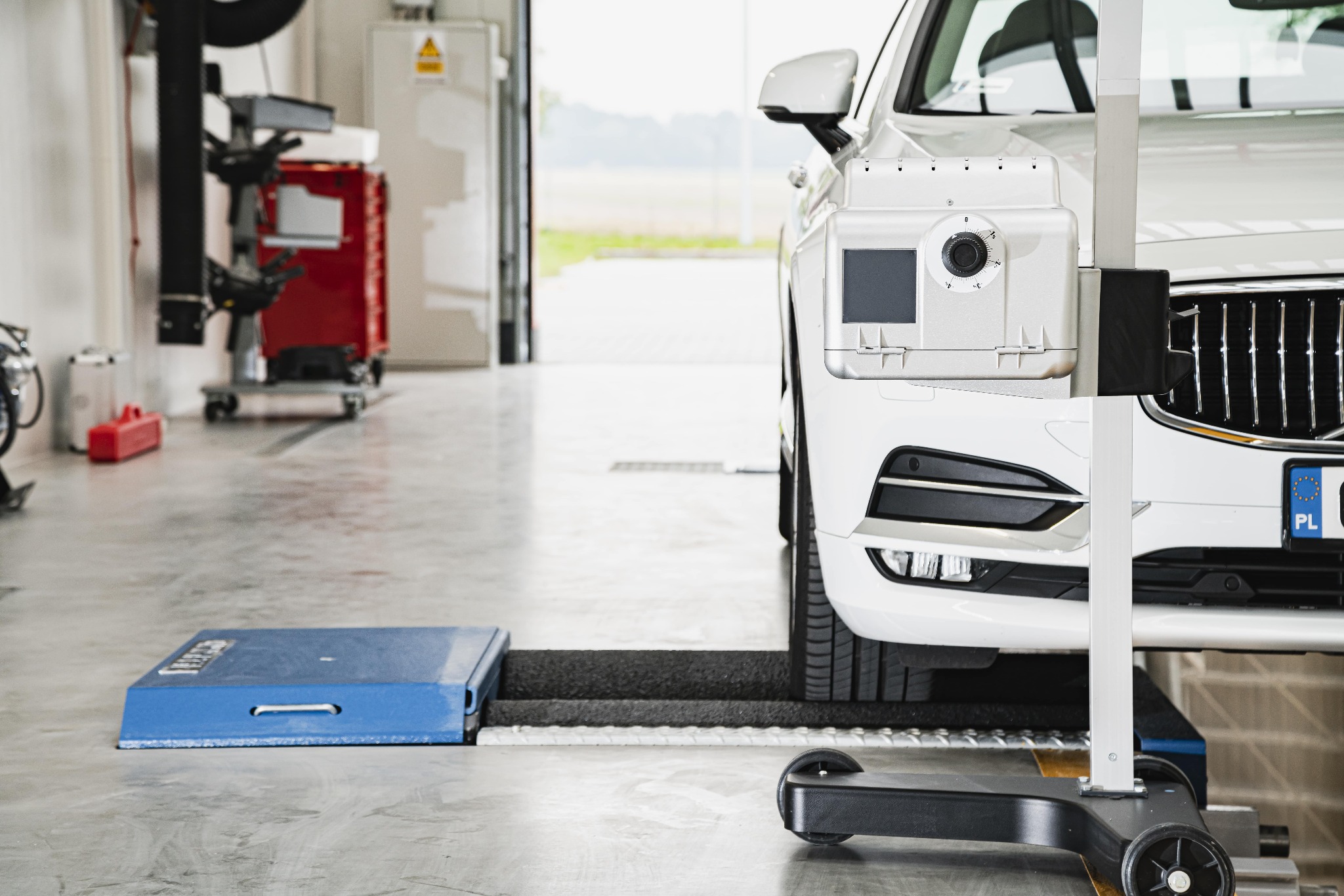 Biały samochód na stanowisku do geometrii kół, z widocznym urządzeniem pomiarowym i płytami najazdowymi w warsztacie.