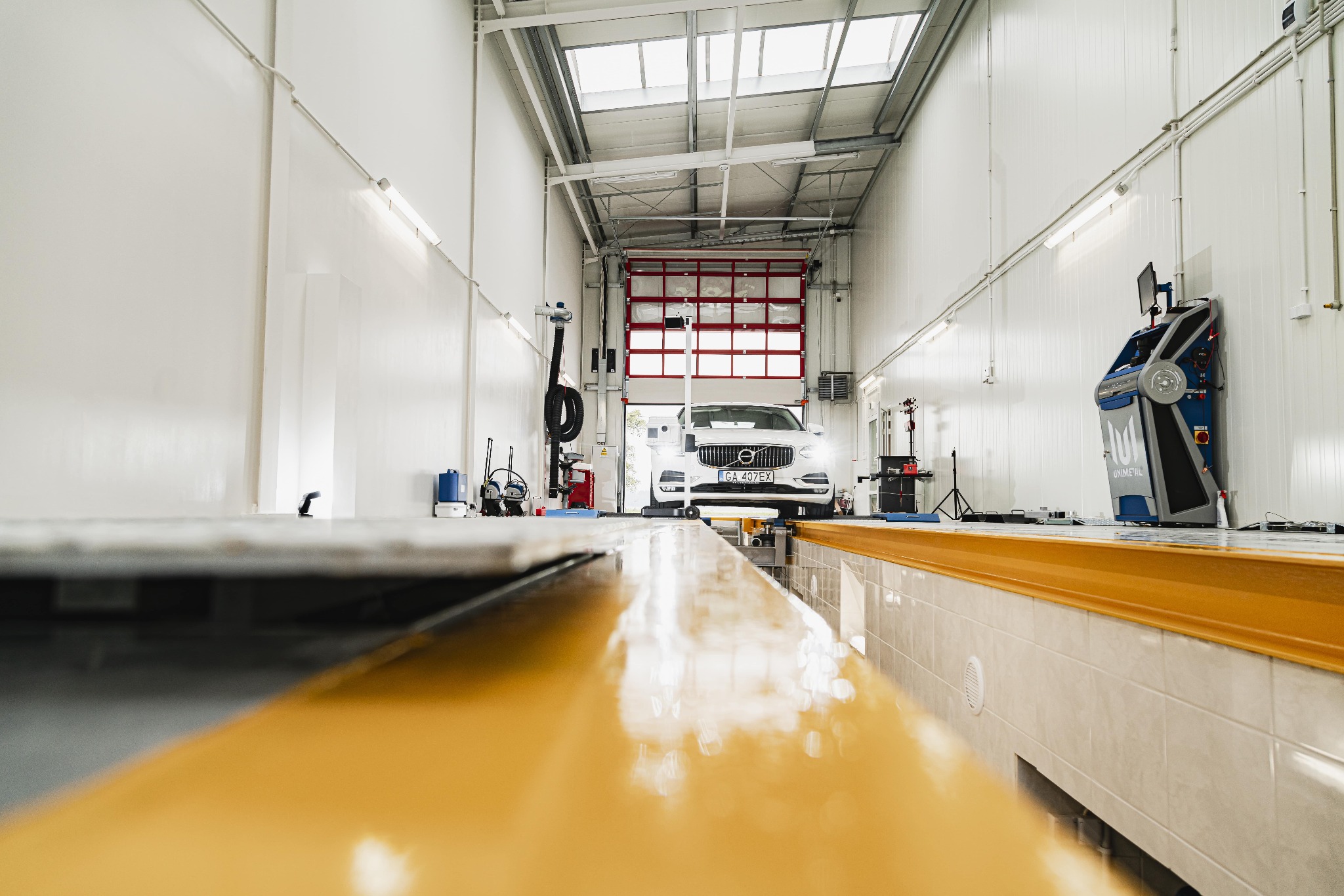 Biały samochód Volvo na podnośniku w jasnym warsztacie z żółtymi elementami, widok z poziomu podnośnika na otwarte drzwi warsztatowe.