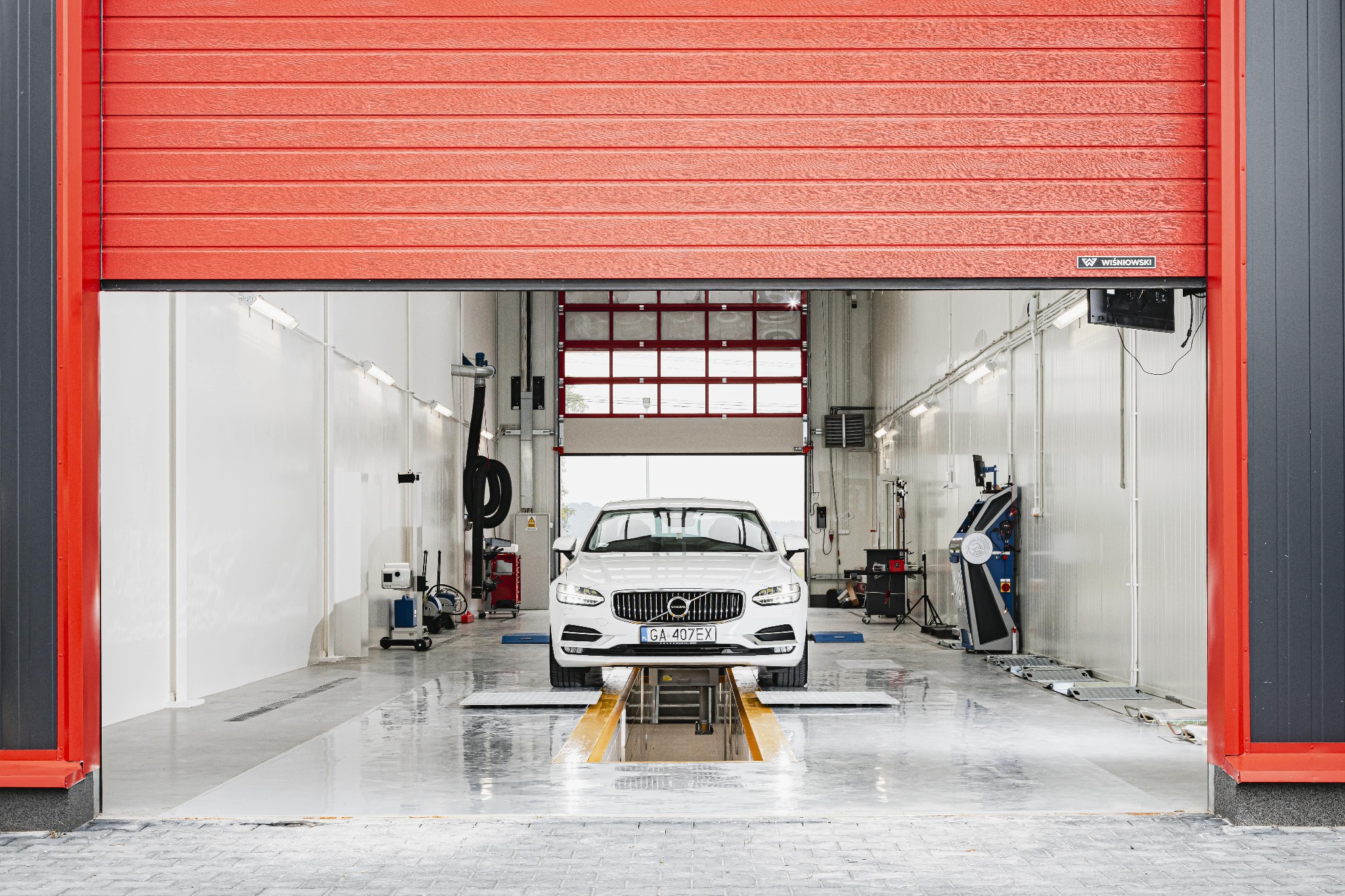 Biały samochód Volvo na podnośniku w jasnym warsztacie samochodowym z częściowo otwartą czerwoną bramą segmentową.