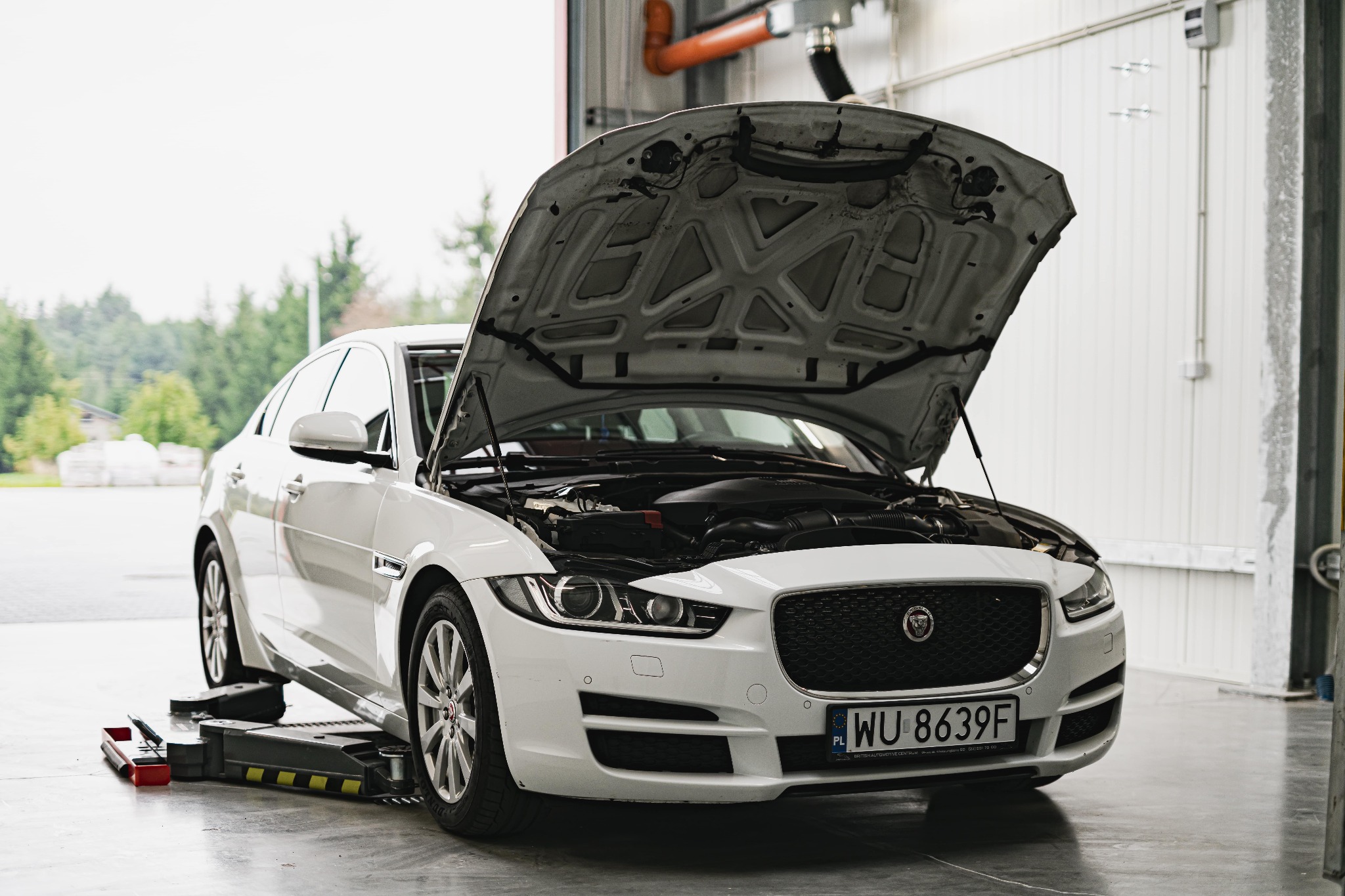 Biały Jaguar XE z otwartą maską, umieszczony na podnośniku w warsztacie, widoczne narzędzia obok