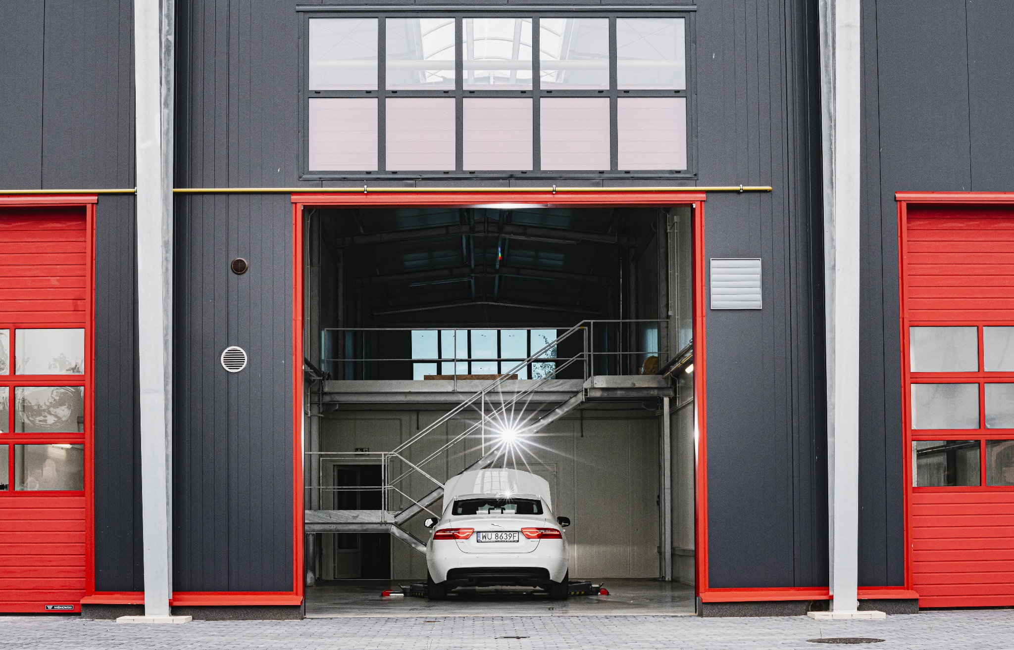 Biały samochód z otwartą maską w nowoczesnym warsztacie z szarą elewacją i czerwonymi bramami.