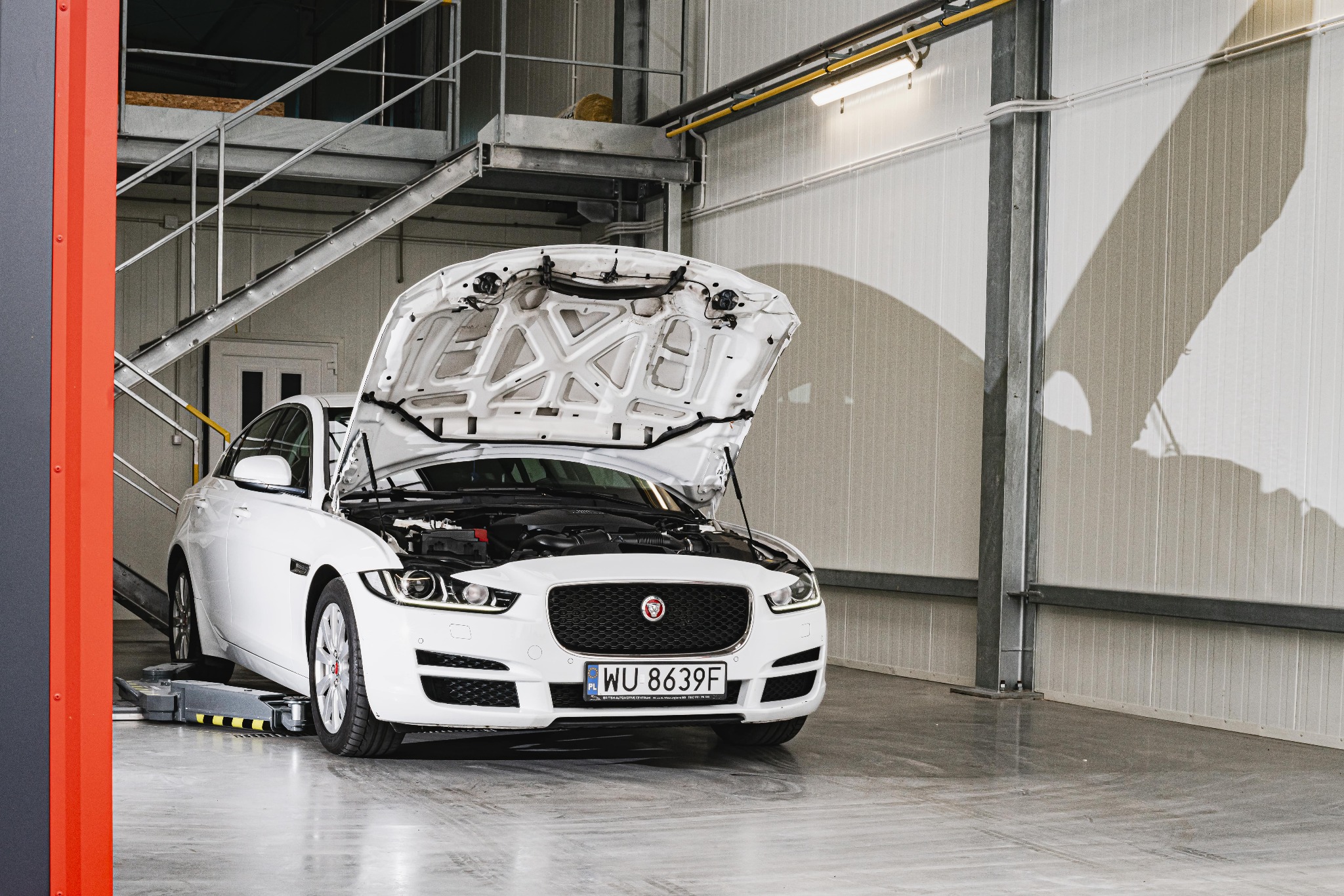 Biały Jaguar XE z otwartą maską, stojący na podnośniku w nowoczesnym warsztacie z metalowymi schodami w tle.