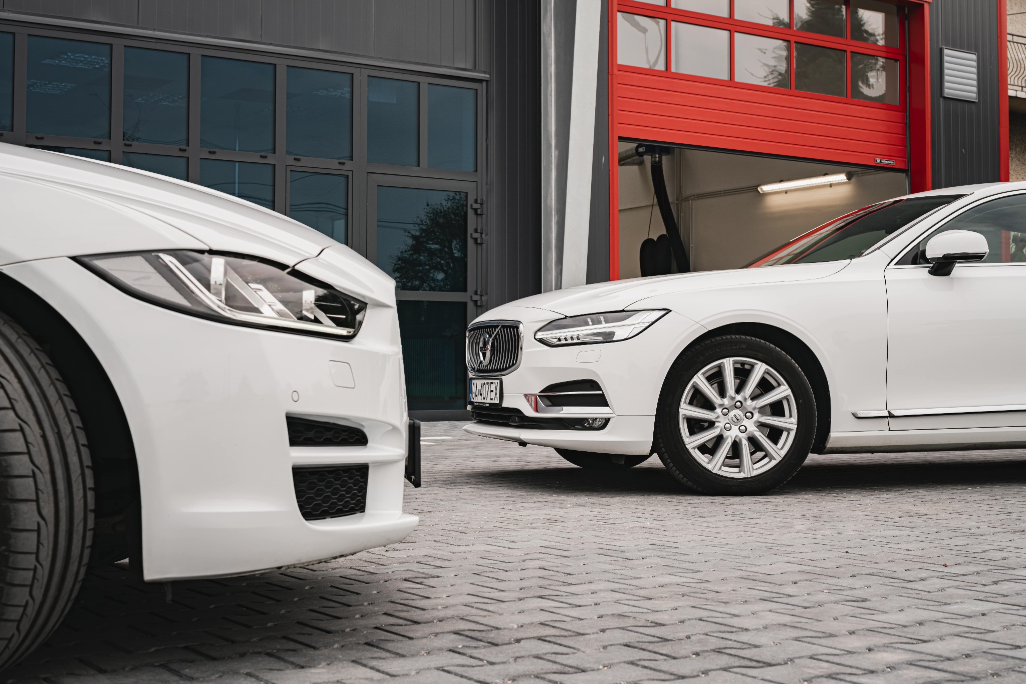 Dwa białe samochody (Jaguar i Volvo) stojące na zewnątrz warsztatu z otwartą czerwoną bramą garażową w tle.