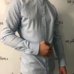 Jasnoniebieska koszula z długim rękawem i monogramem 'JD' na mankiecie, zapinana na białe guziki, widoczna na tle z logotypem.