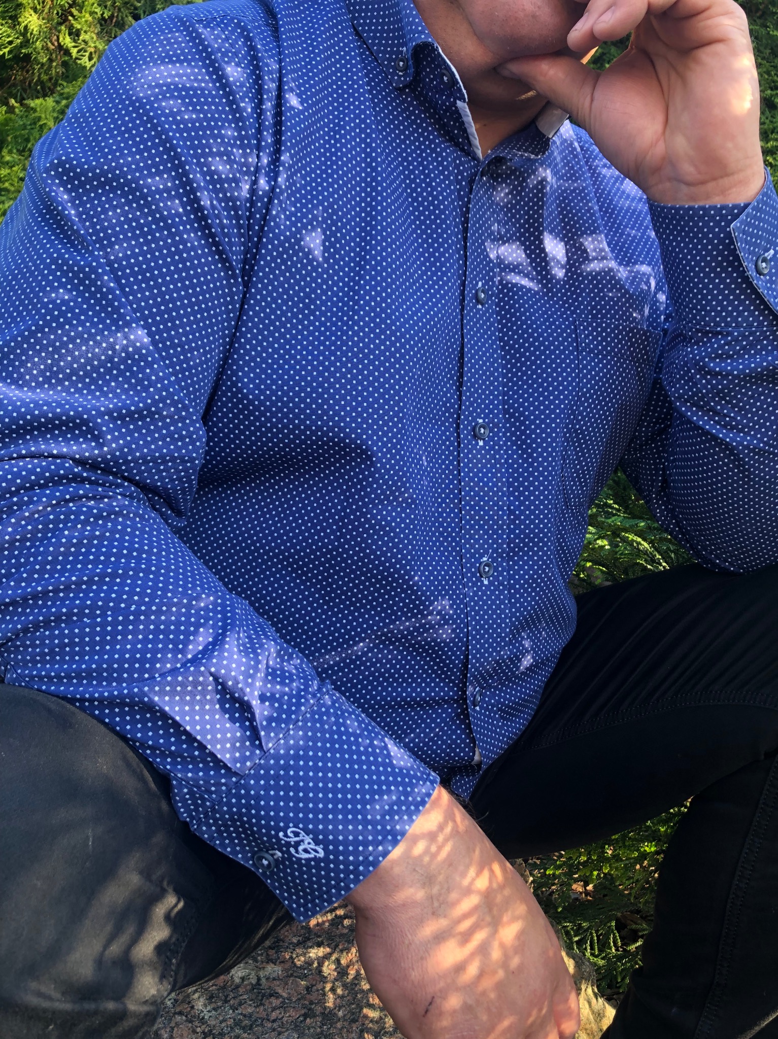 Mężczyzna w niebieskiej koszuli w białe kropki siedzi na zewnątrz, z ręką na brodzie, z widocznym monogramem na mankiecie.