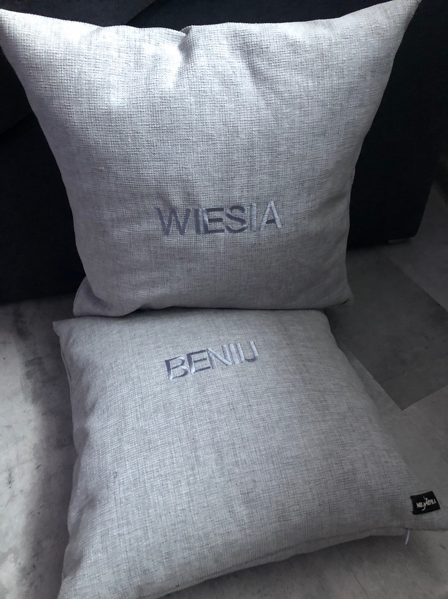 Dwie lniane poduszki dekoracyjne z wyhaftowanymi imionami: 'Wiesia' na górnej i 'Benia' na dolnej, ustawione na podłodze.