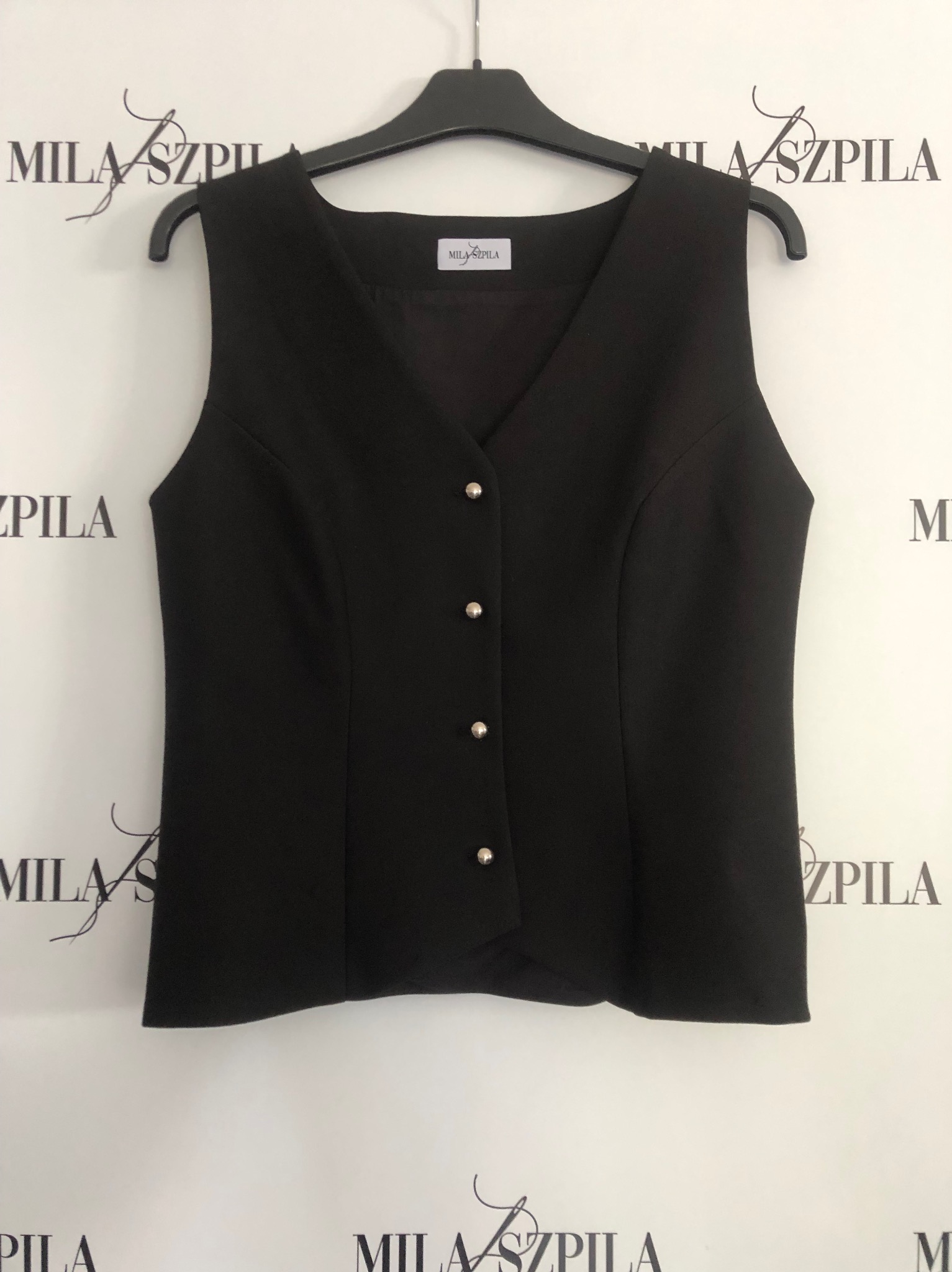 Czarna, elegancka kamizelka damska zapinana na cztery srebrne guziki, wisząca na czarnym wieszaku na tle ściany z powtarzającym się logo marki.