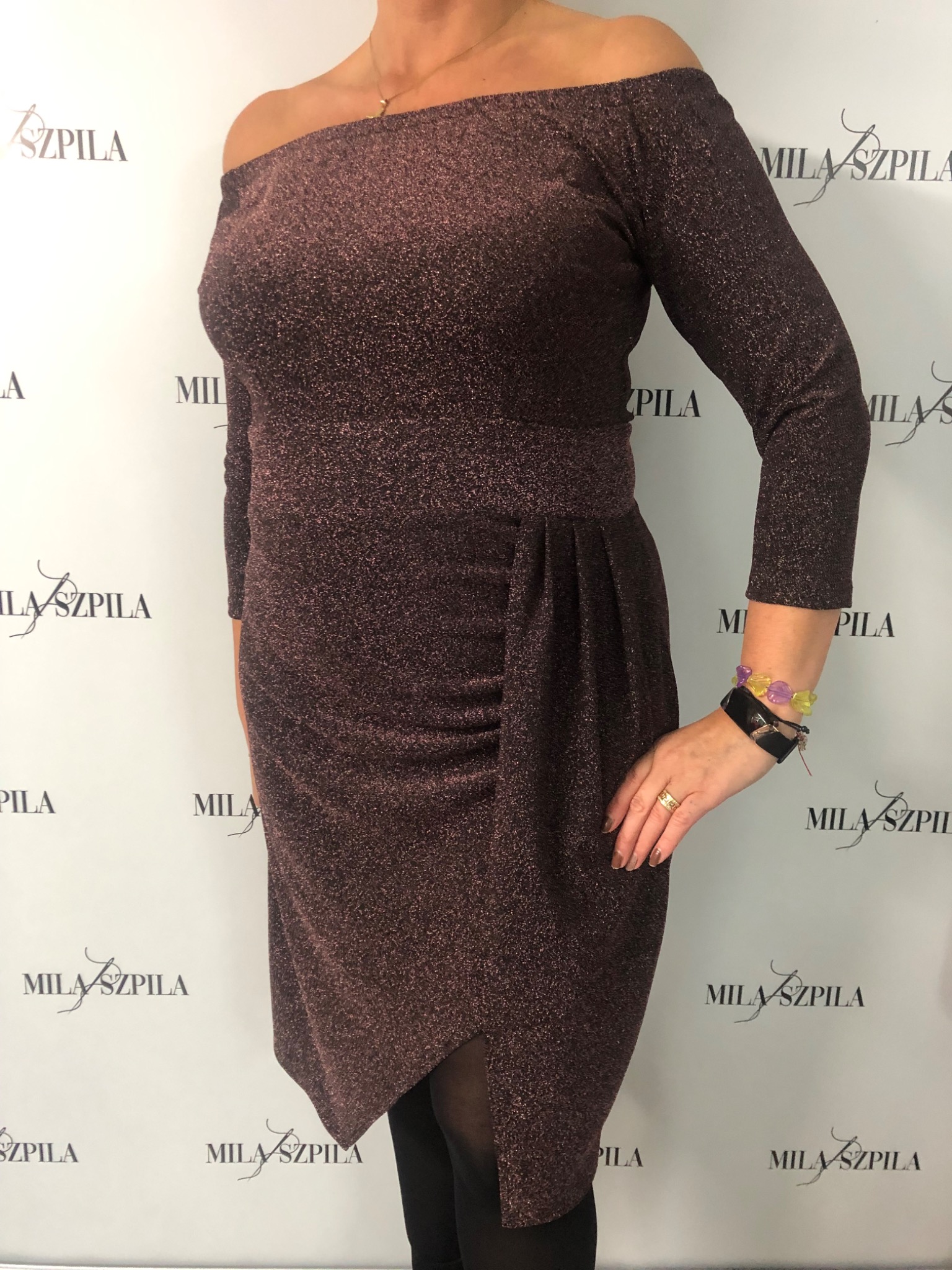 Kobieta prezentuje błyszczącą, bordową sukienkę z odkrytymi ramionami, z marszczeniem na biodrze i asymetrycznym dołem, pozując na tle z powtarzającym się logo.