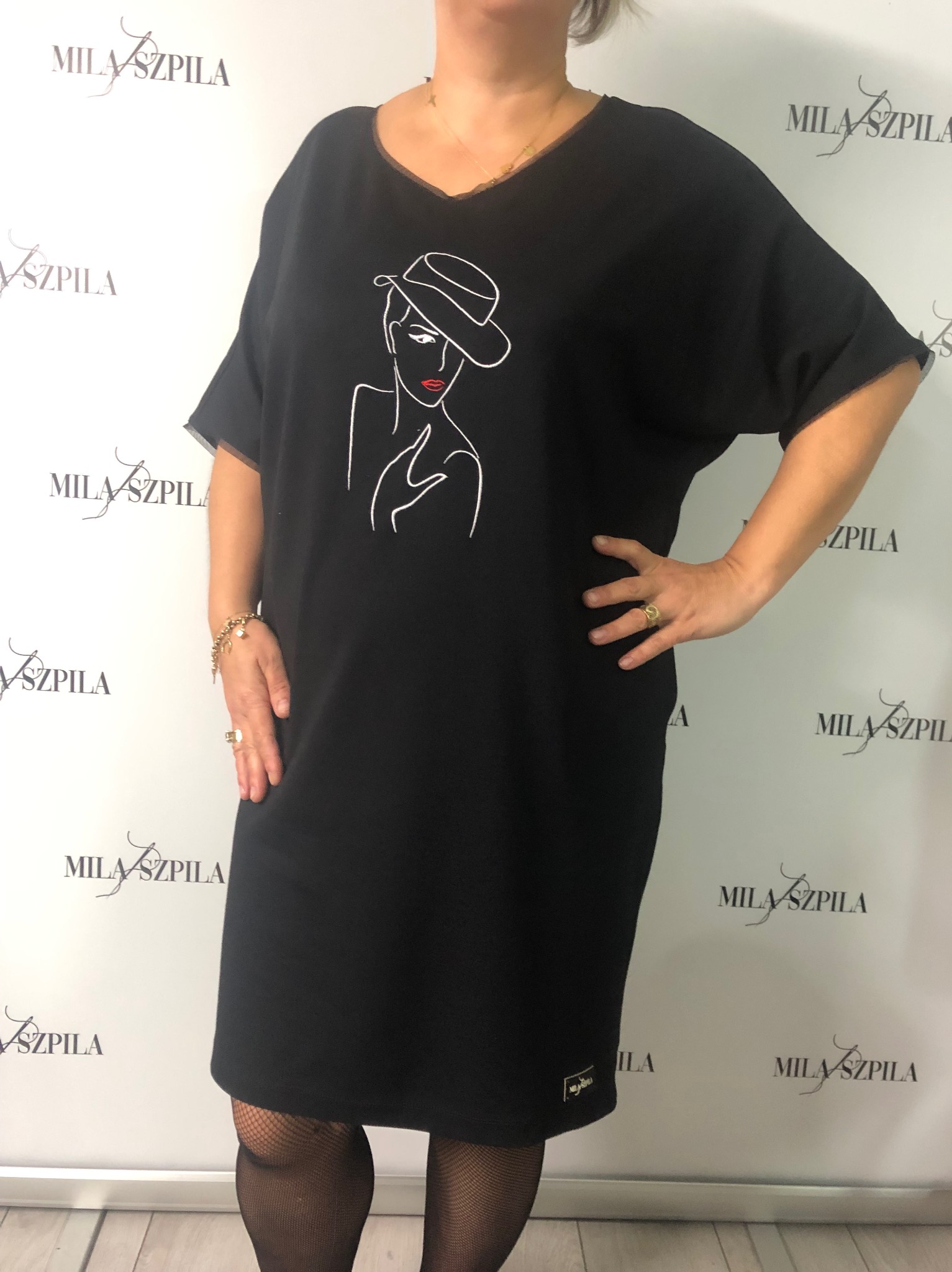 Kobieta prezentuje czarną sukienkę z krótkim rękawem i graficznym nadrukiem kobiety w kapeluszu, pozuje na tle z powtarzającym się logo marki.